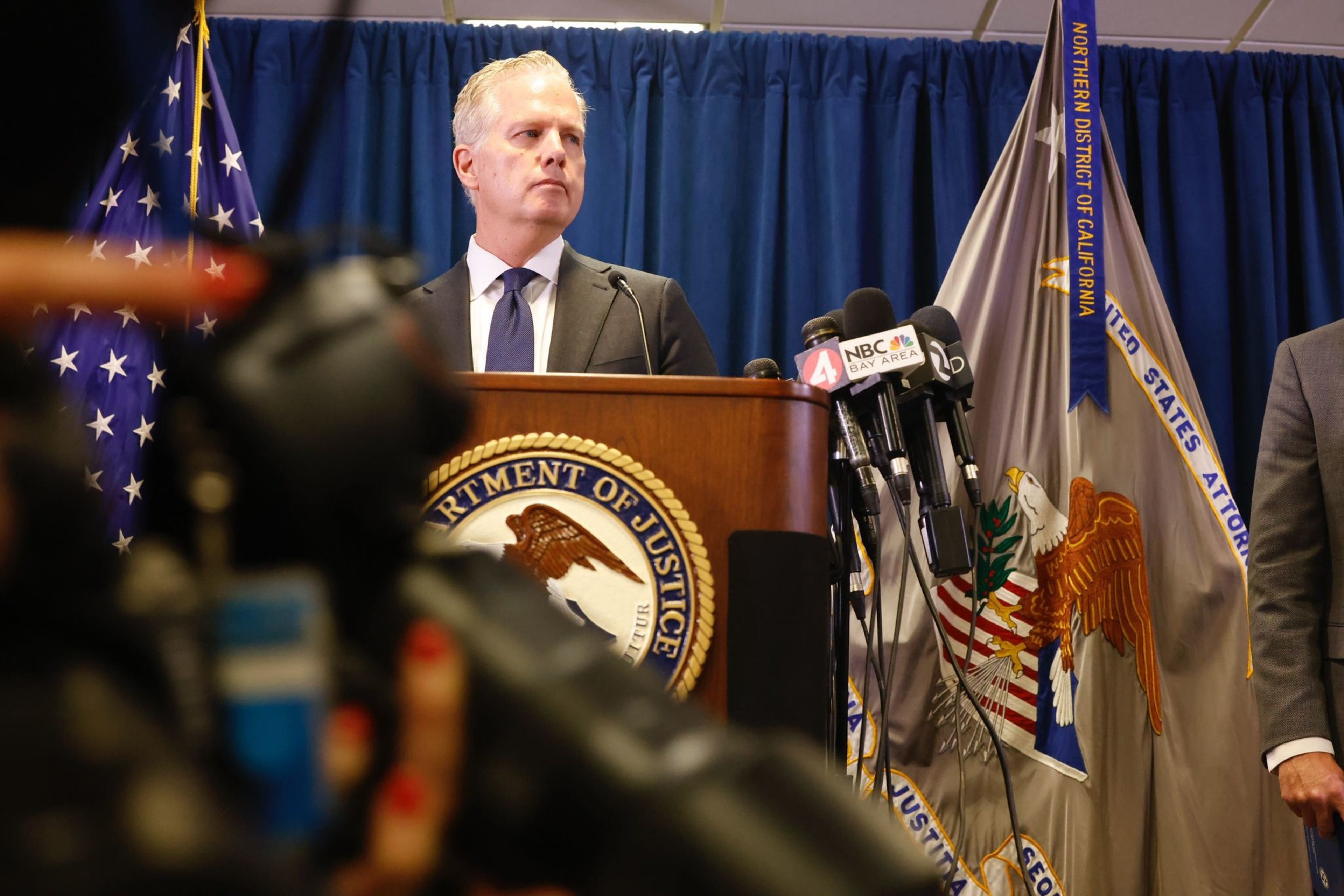 A man in a suit stands at a podium with "Department of Justice" signage, surrounded by flags and microphones from several news outlets.
