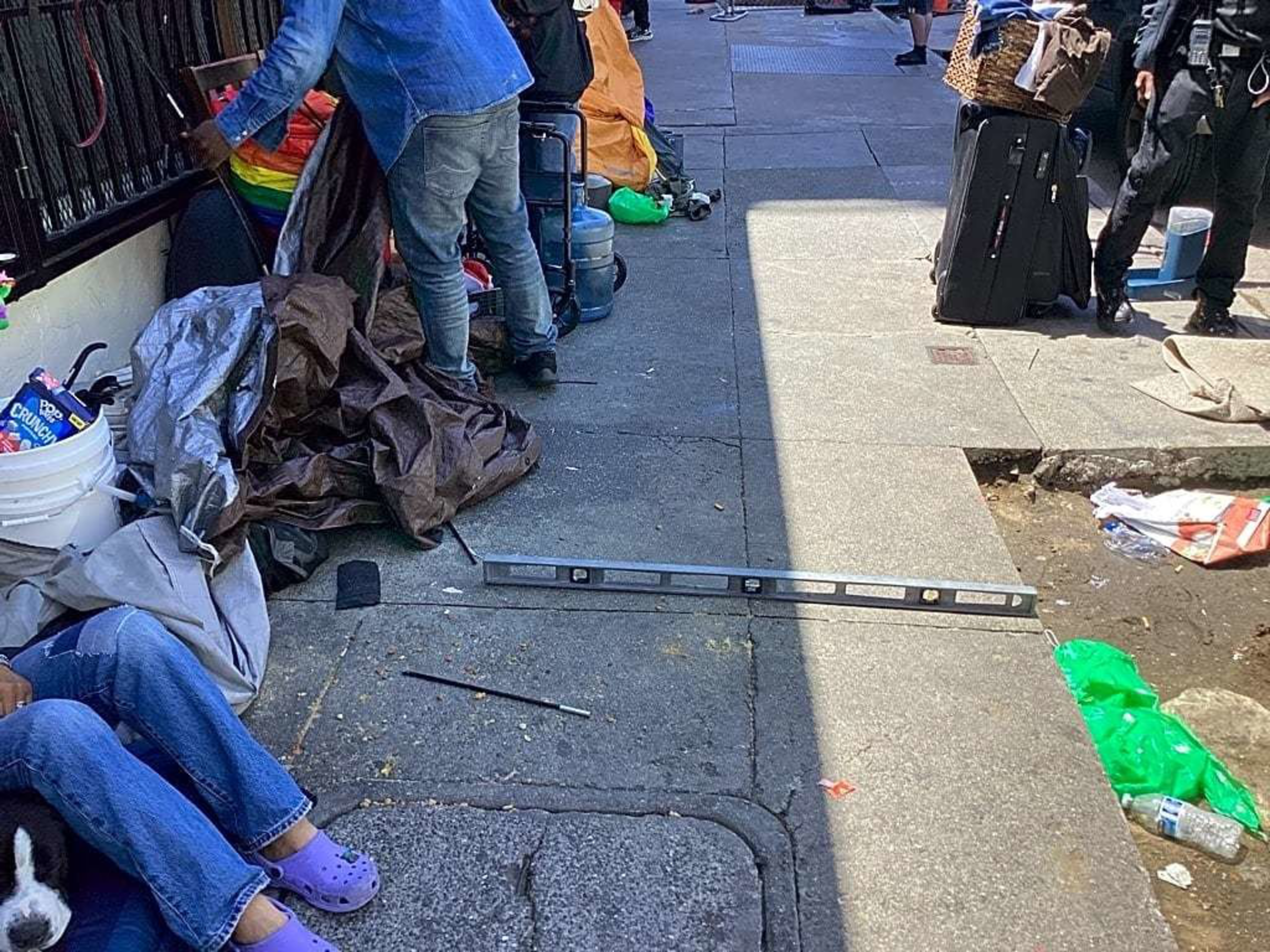 A bustling sidewalk scene shows people with belongings, including luggage and bags. One person is seated with a dog, and various items like tarps and tools are scattered around.