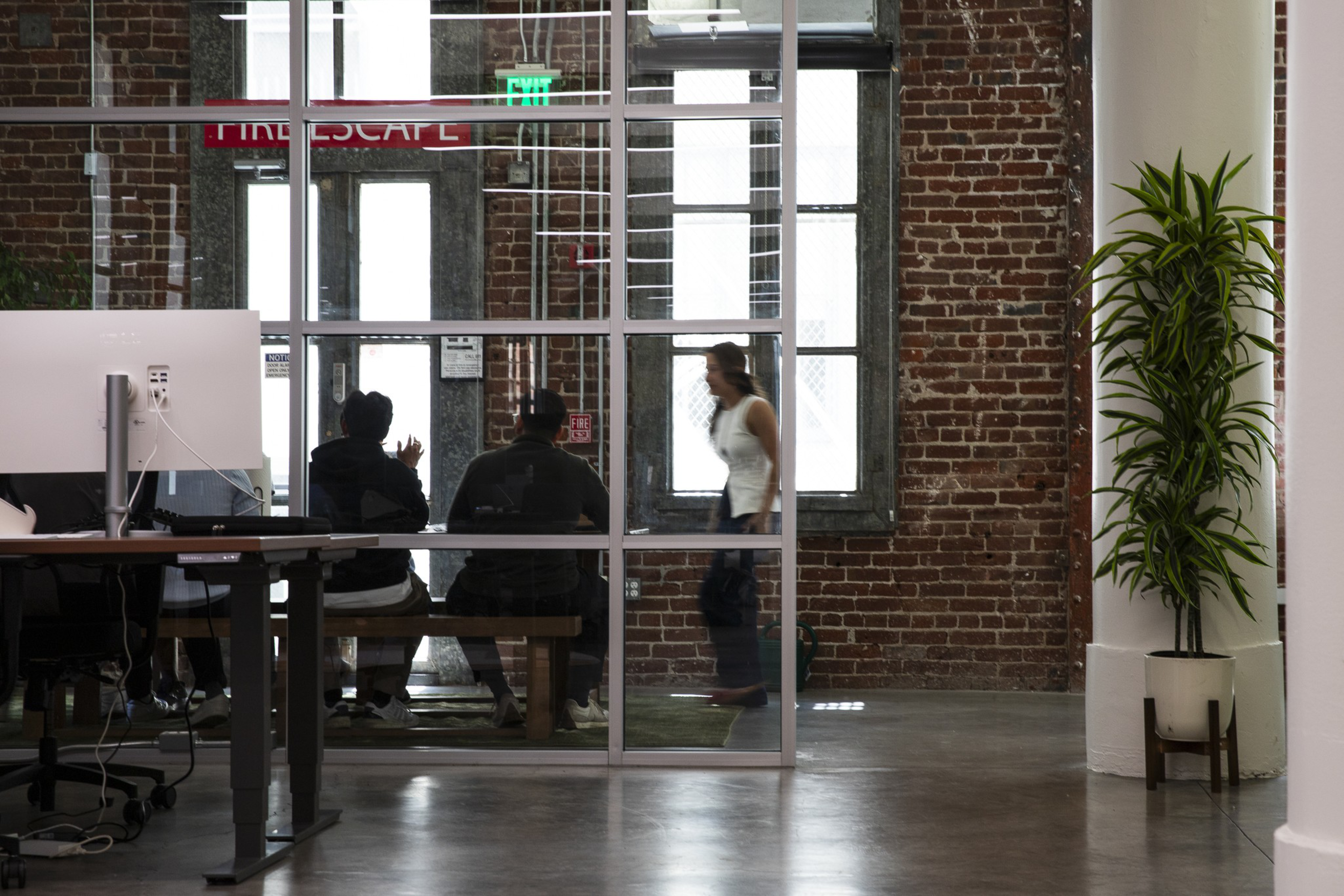 Three people sit inside a glass-walled room with exposed brick walls, while a woman walks past outside, and a plant stands near a white pillar.