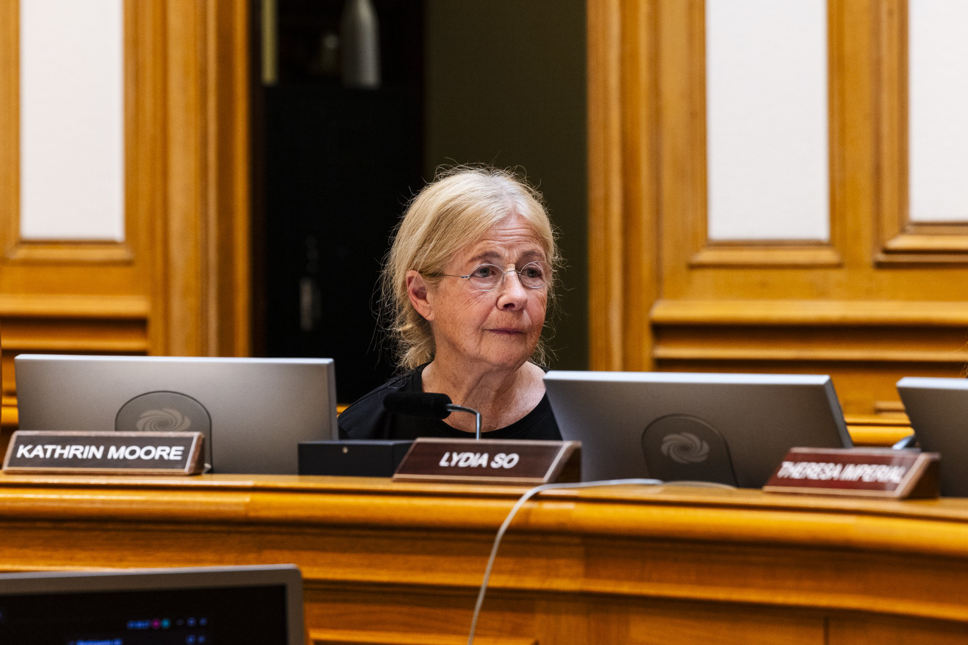 An older woman with glasses sits at a wooden desk with multiple nameplates and monitors in front of her in a formal setting, possibly a government meeting.