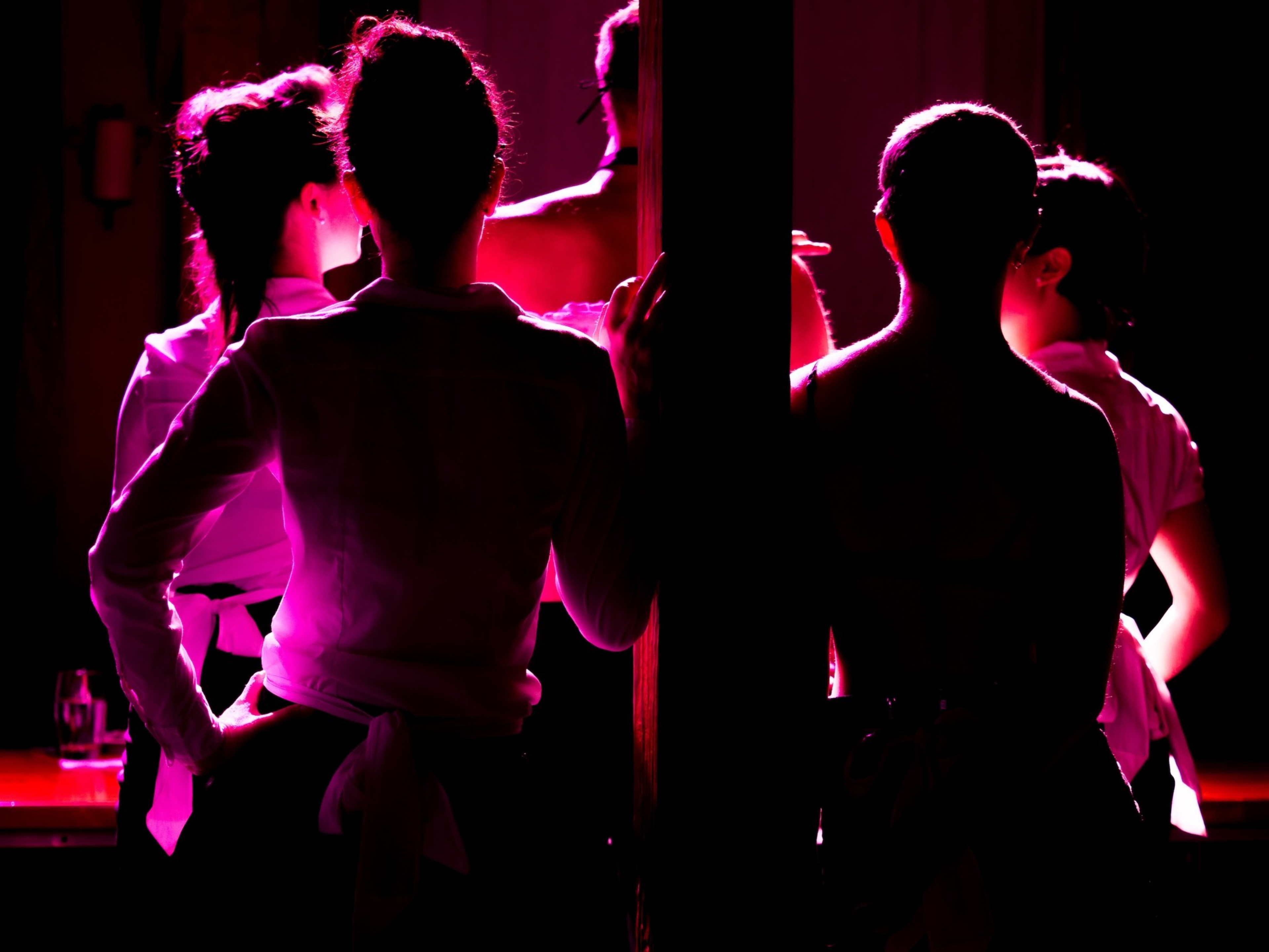 Five people are gathered in dim red and pink lighting, with most facing away and one partially turned, creating a silhouetted effect.