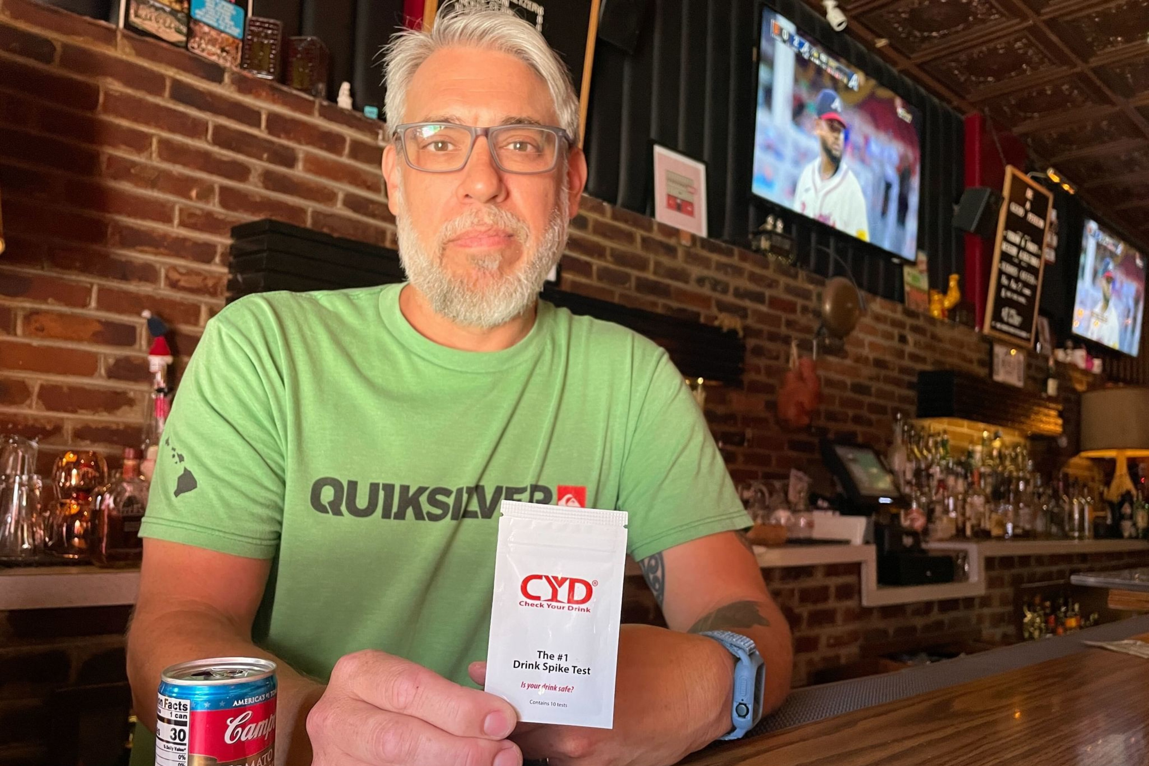 A man stands behind a bar holding a white package that says "Drink Spike Test" on the front.