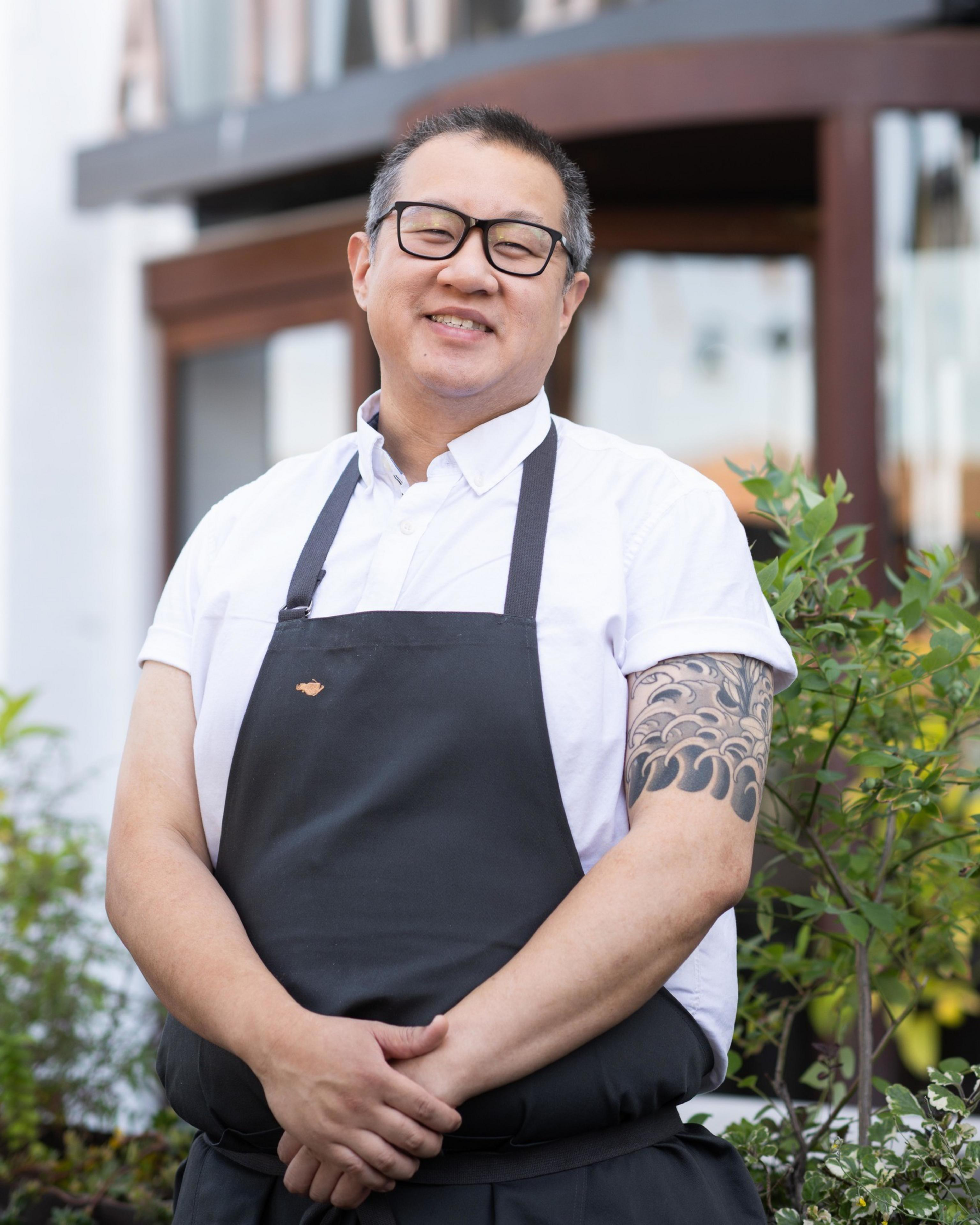 A person stands outside, smiling, wearing glasses, a white short-sleeve shirt, and a dark apron. They have a tattoo on their left arm and are in a garden setting.
