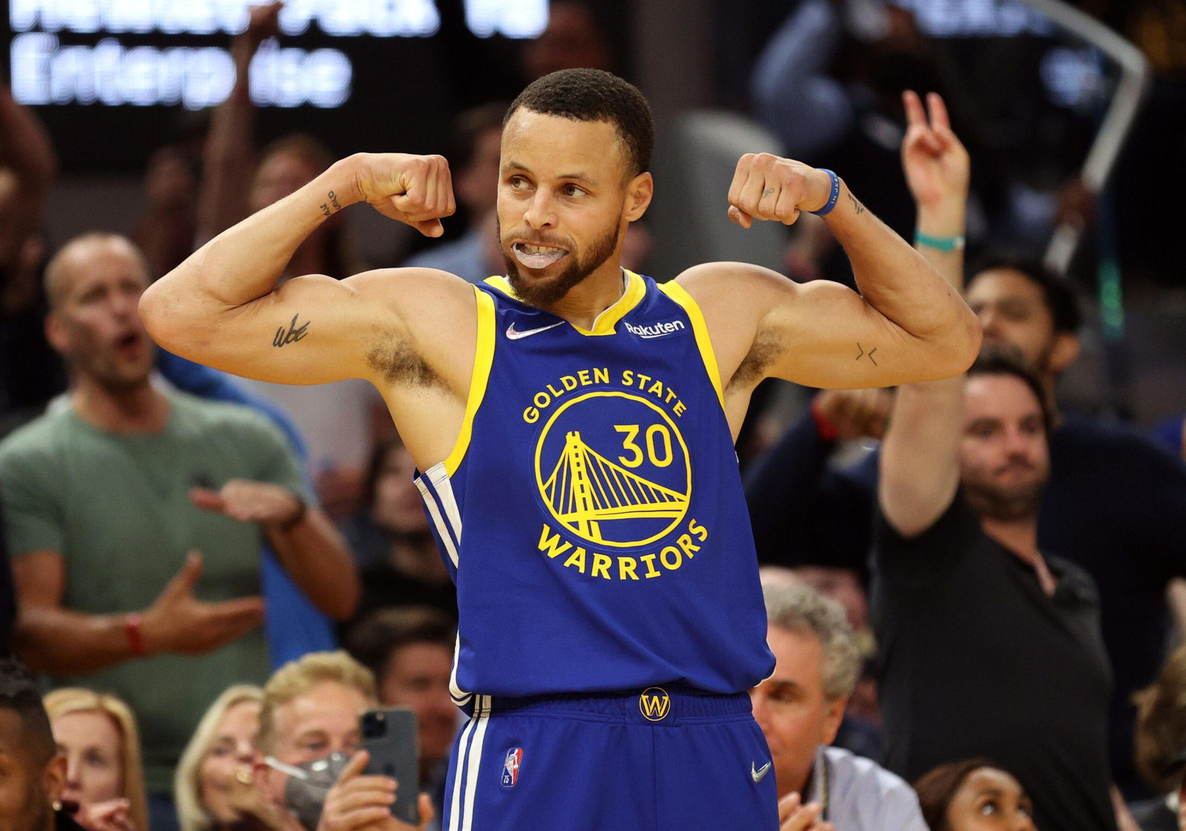 A basketball player in a Golden State Warriors jersey flexes both arms, showing his muscles while fans cheer in the background.