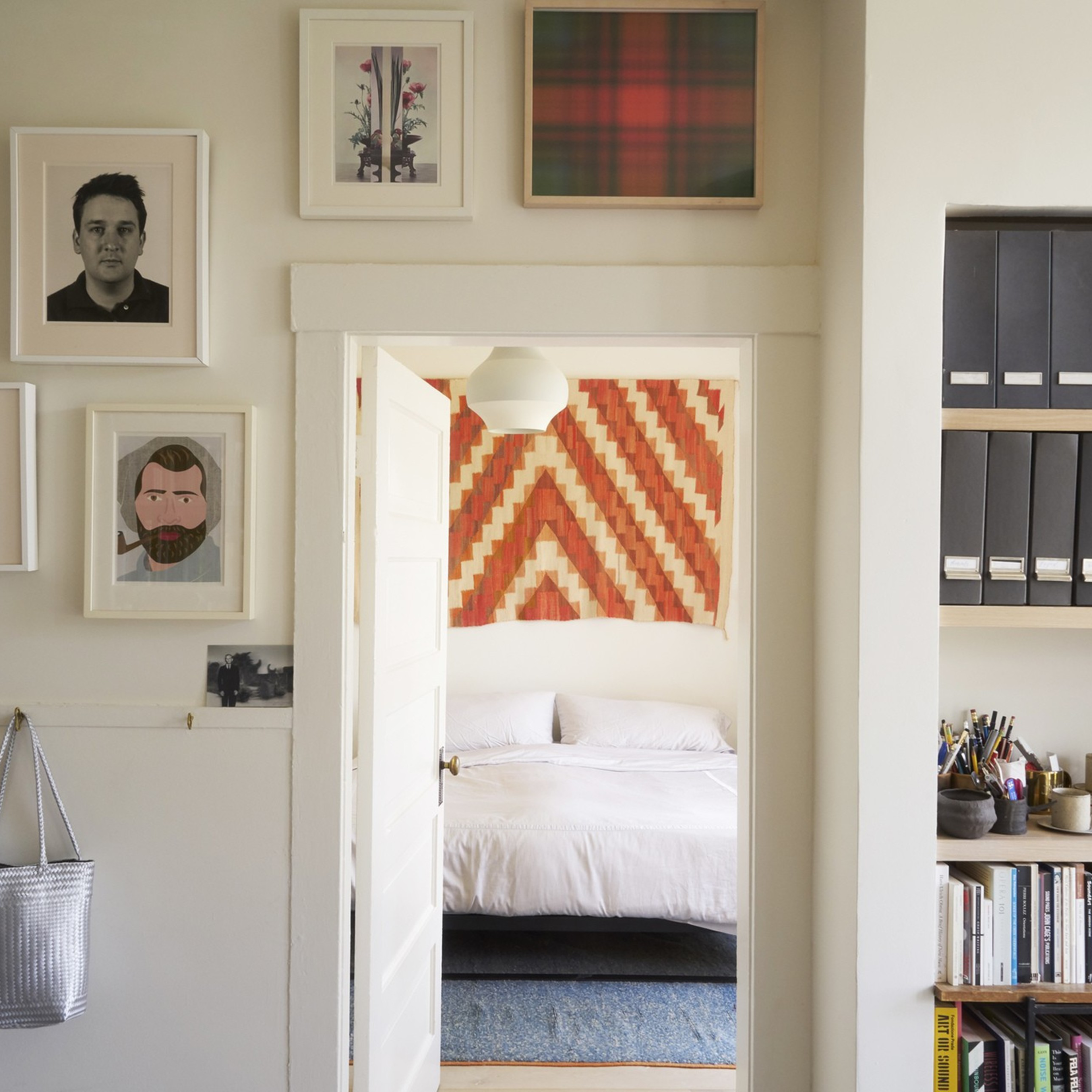 A cozy room with art on the walls, a door leading to a bedroom with a patterned decoration, shelves with files and books, and bags hanging on the walls.