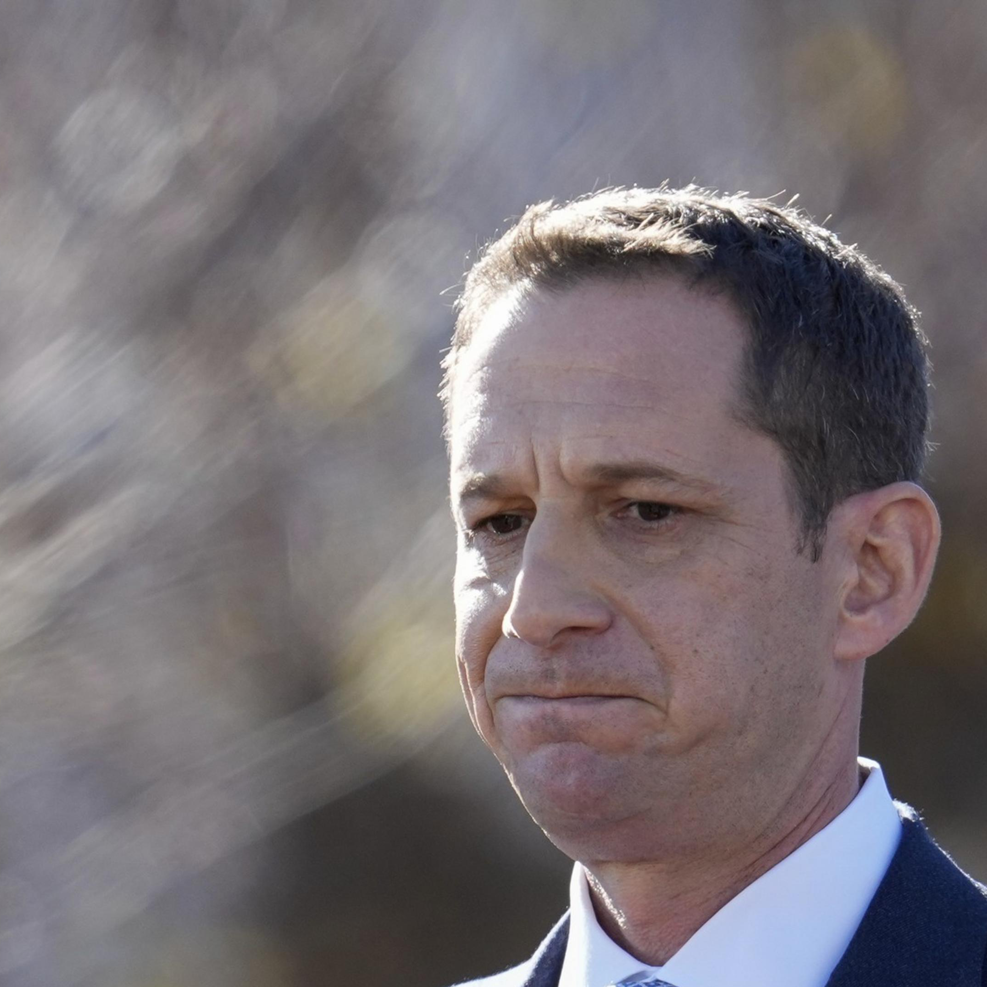 A man in a dark suit and light blue tie gazes pensively, set against a blurred outdoor background with soft lighting.