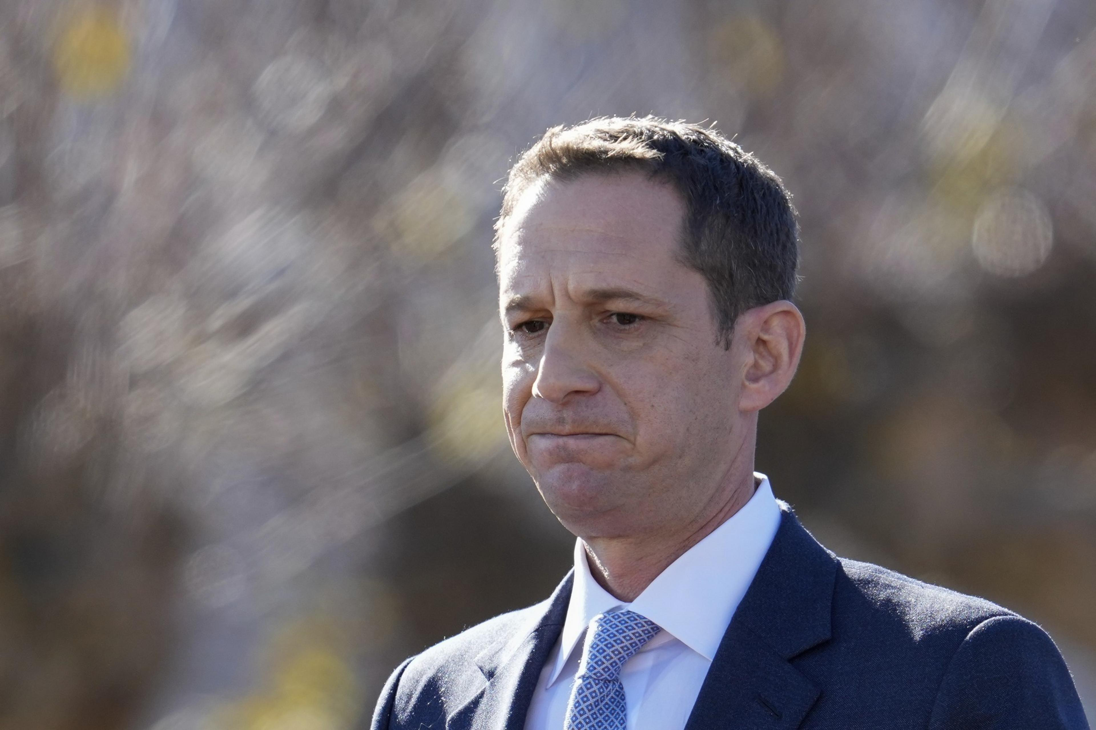 A man in a dark suit and light blue tie gazes pensively, set against a blurred outdoor background with soft lighting.