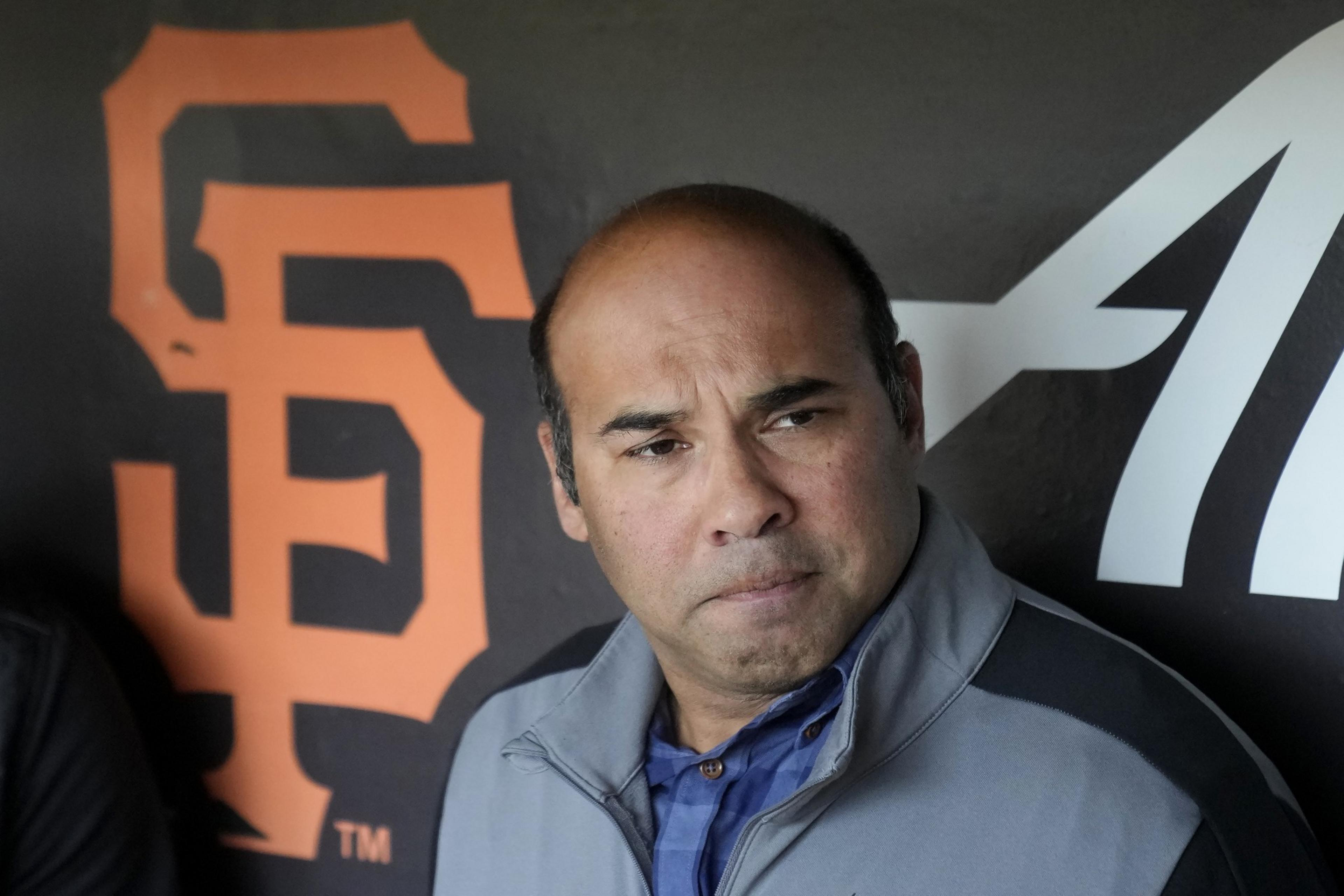 A man in a gray jacket and blue checked shirt is standing in front of a wall adorned with the orange San Francisco Giants logo. He has a serious expression.