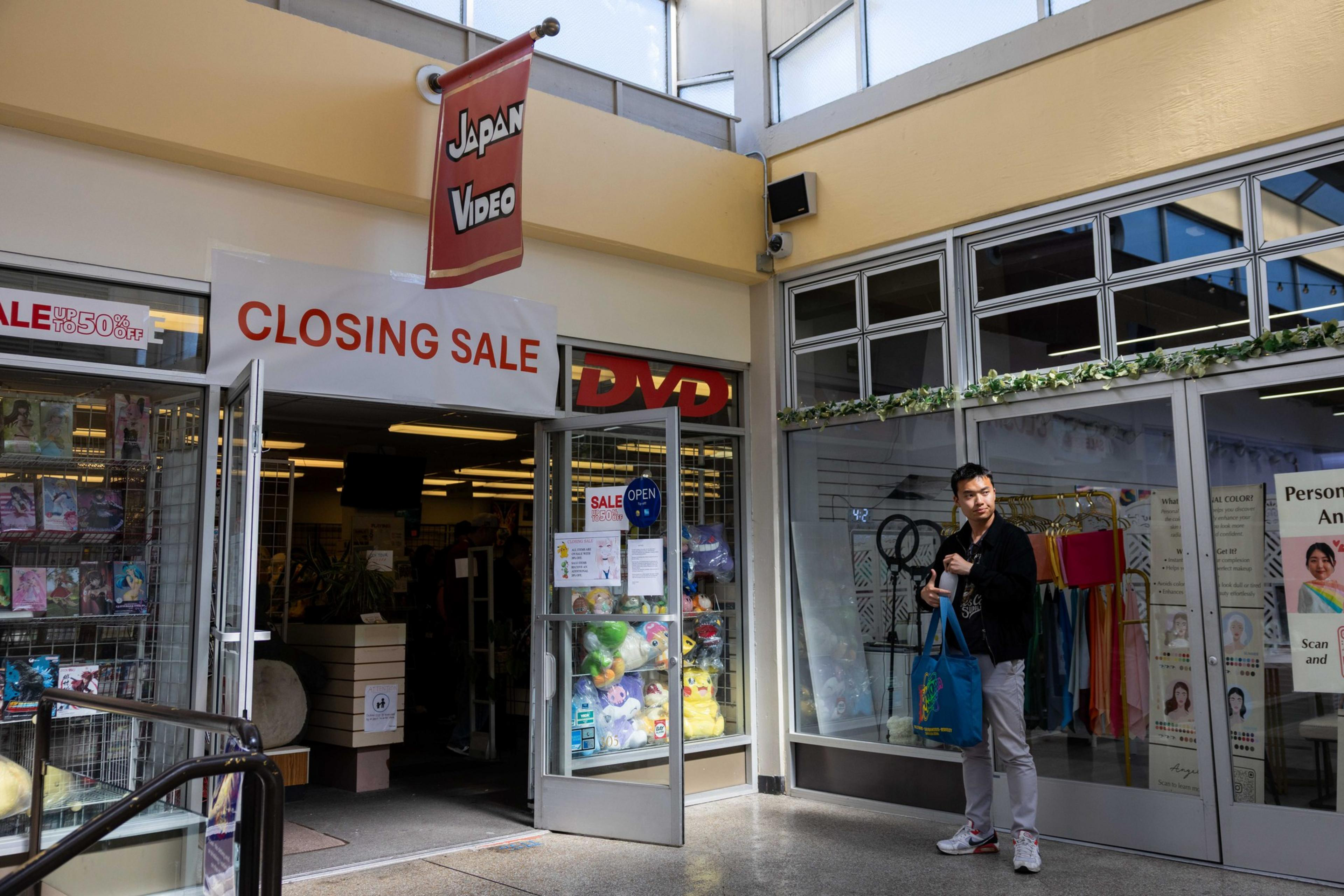 A store with a &quot;Closing Sale&quot; sign features DVDs and plush toys in its entrance. A person holding a blue bag stands outside, next to another shop with colorful items.