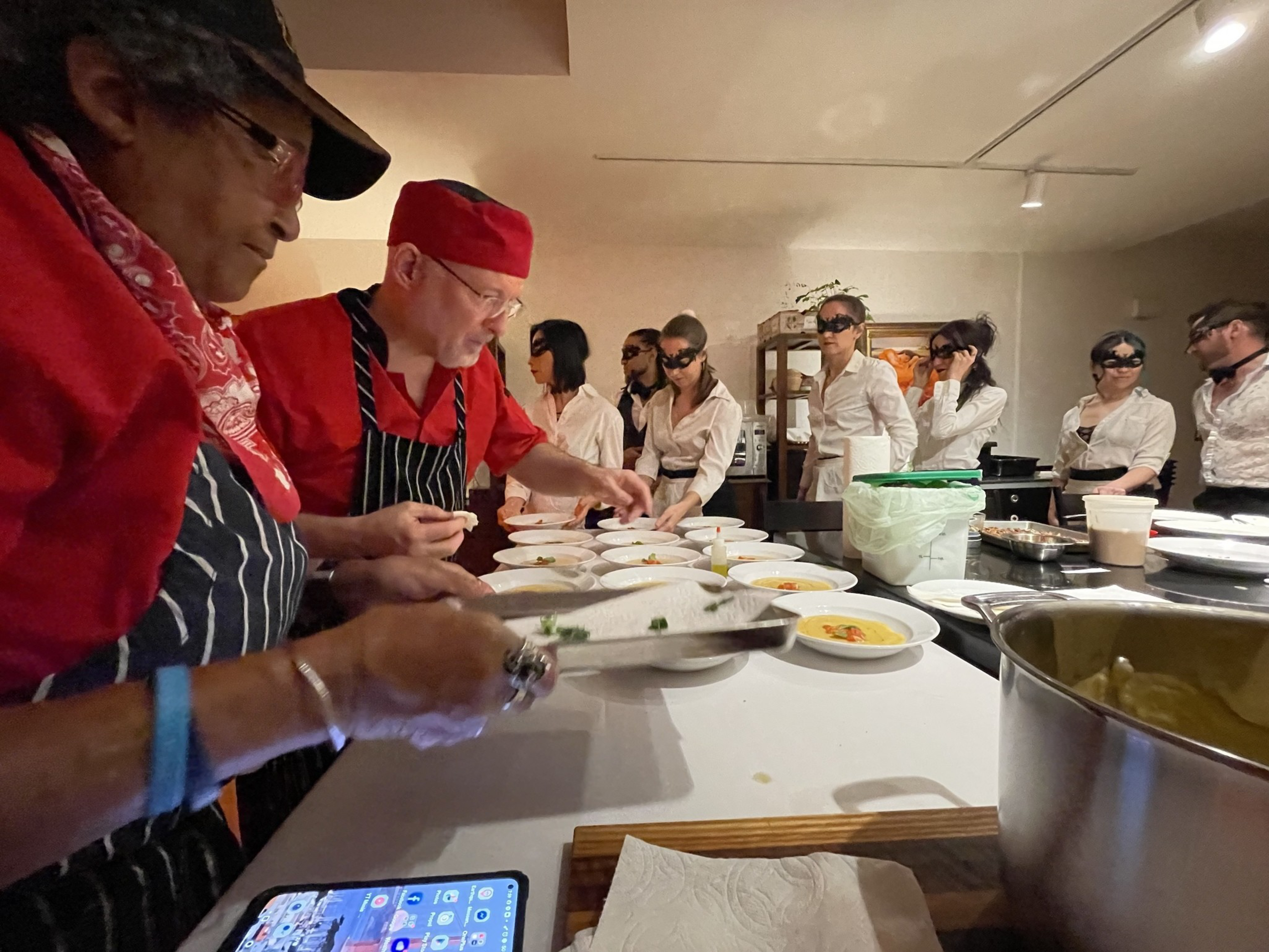 Two chefs in red and black prepare dishes while people in white shirts and black eye masks stand in line on the other side of the table.