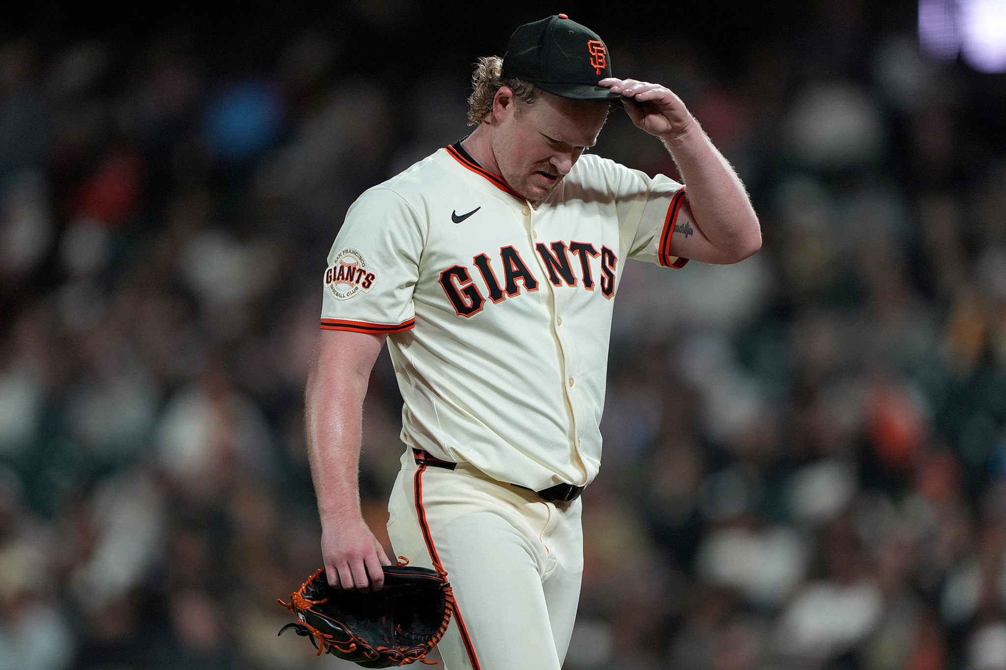 A baseball player in a San Francisco Giants uniform looks down while adjusting his cap, holding a glove in his other hand on a dimly lit field.