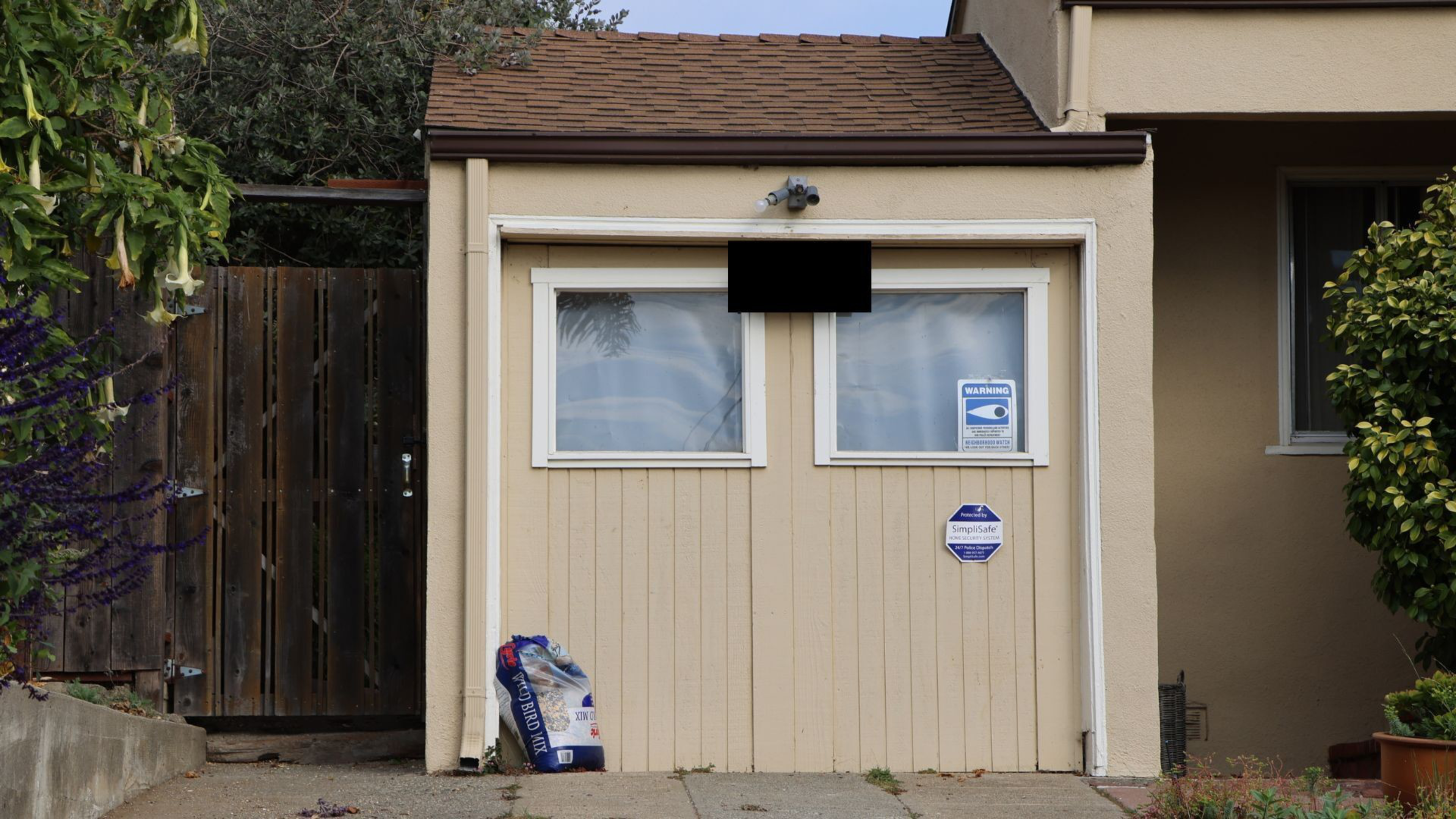 A beige garage door with two upper windows, security cameras above, warning and alarm system signs, a blue bag on the ground, and surrounded by plants and a wooden fence.