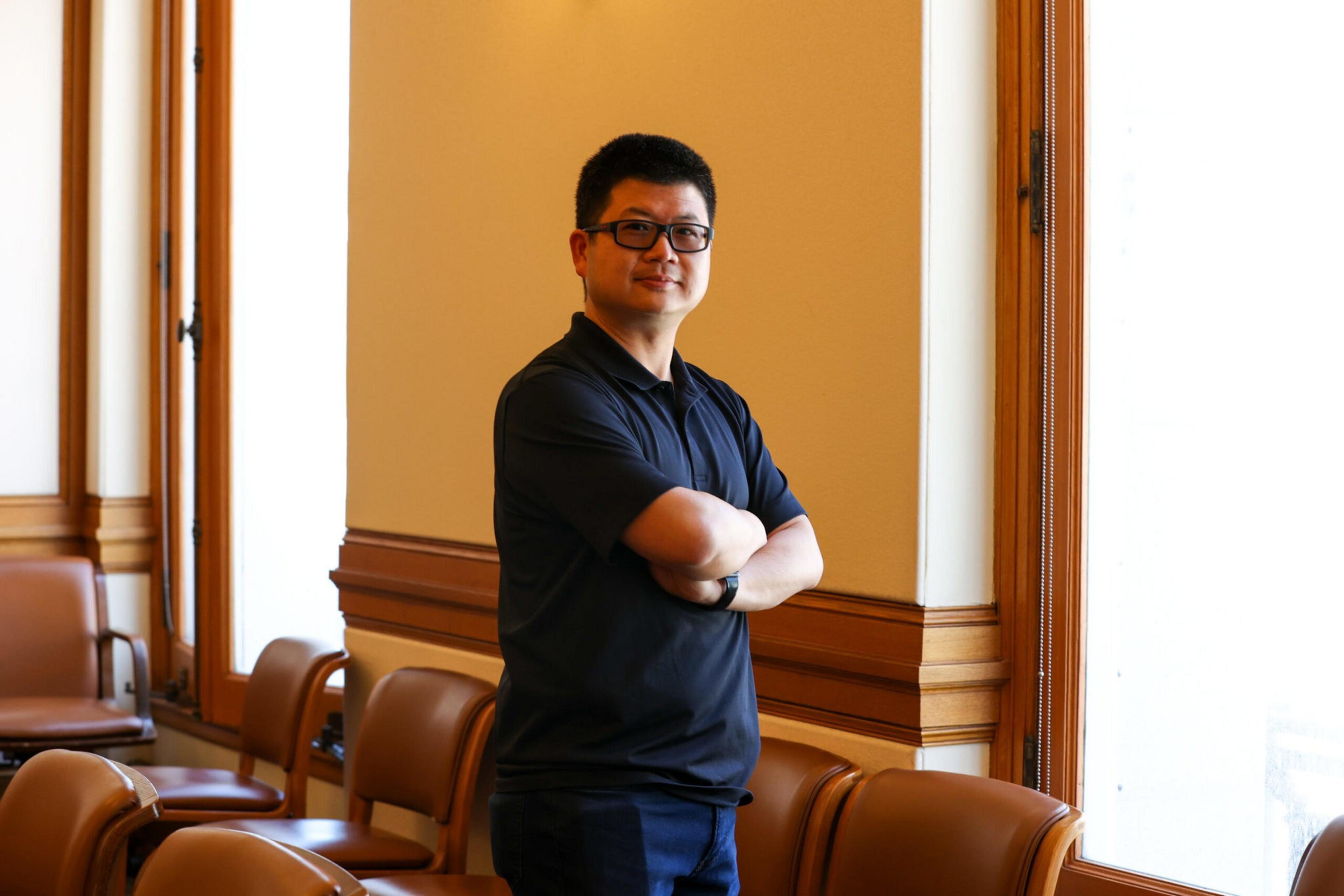 A man wearing glasses and a black polo shirt stands with arms crossed in a room with wooden paneling and brown chairs by large windows.