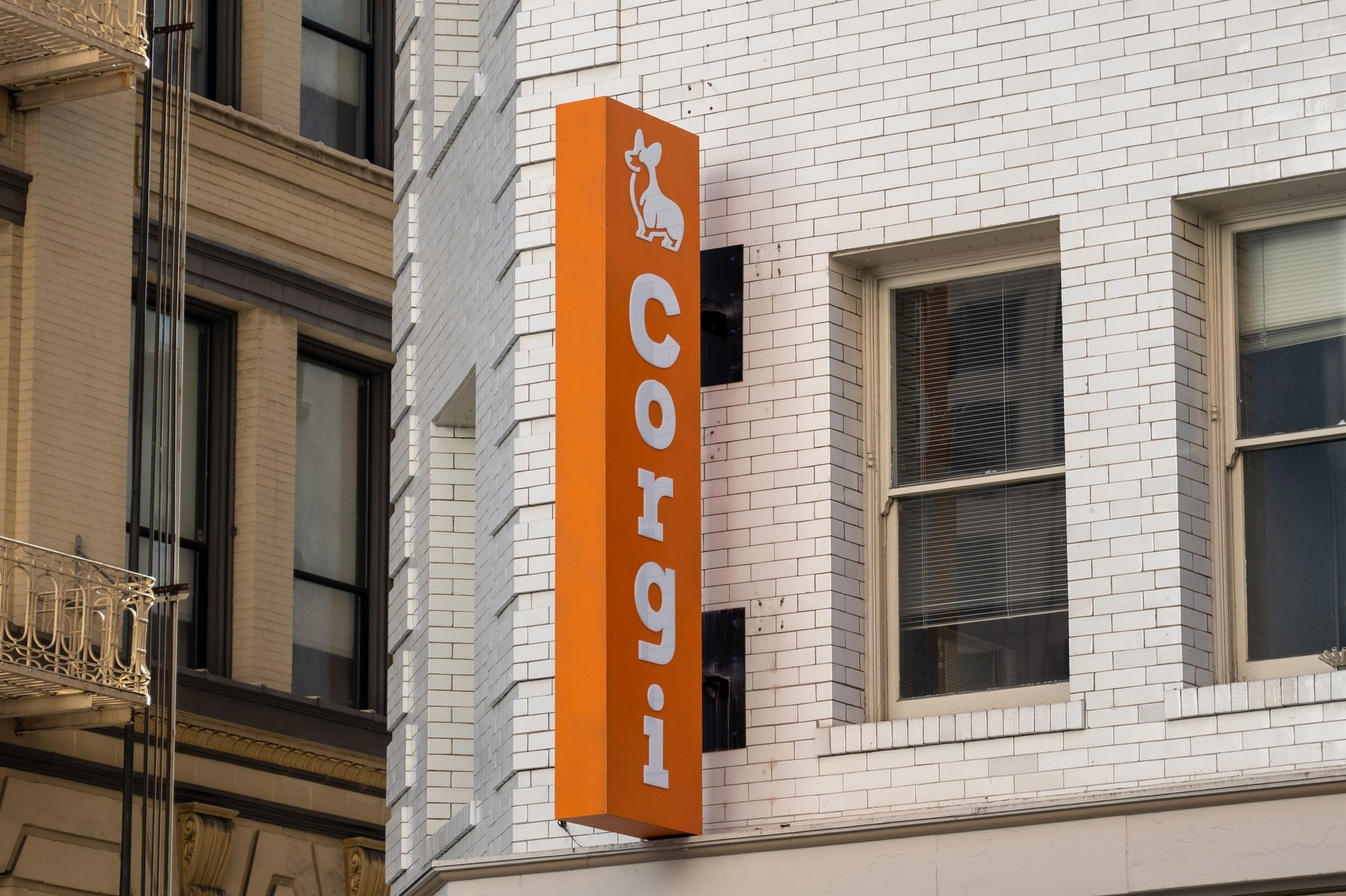 An orange vertical sign with a white corgi dog silhouette and the word &acirc;Corgi&acirc; in white letters is mounted on a white brick building.