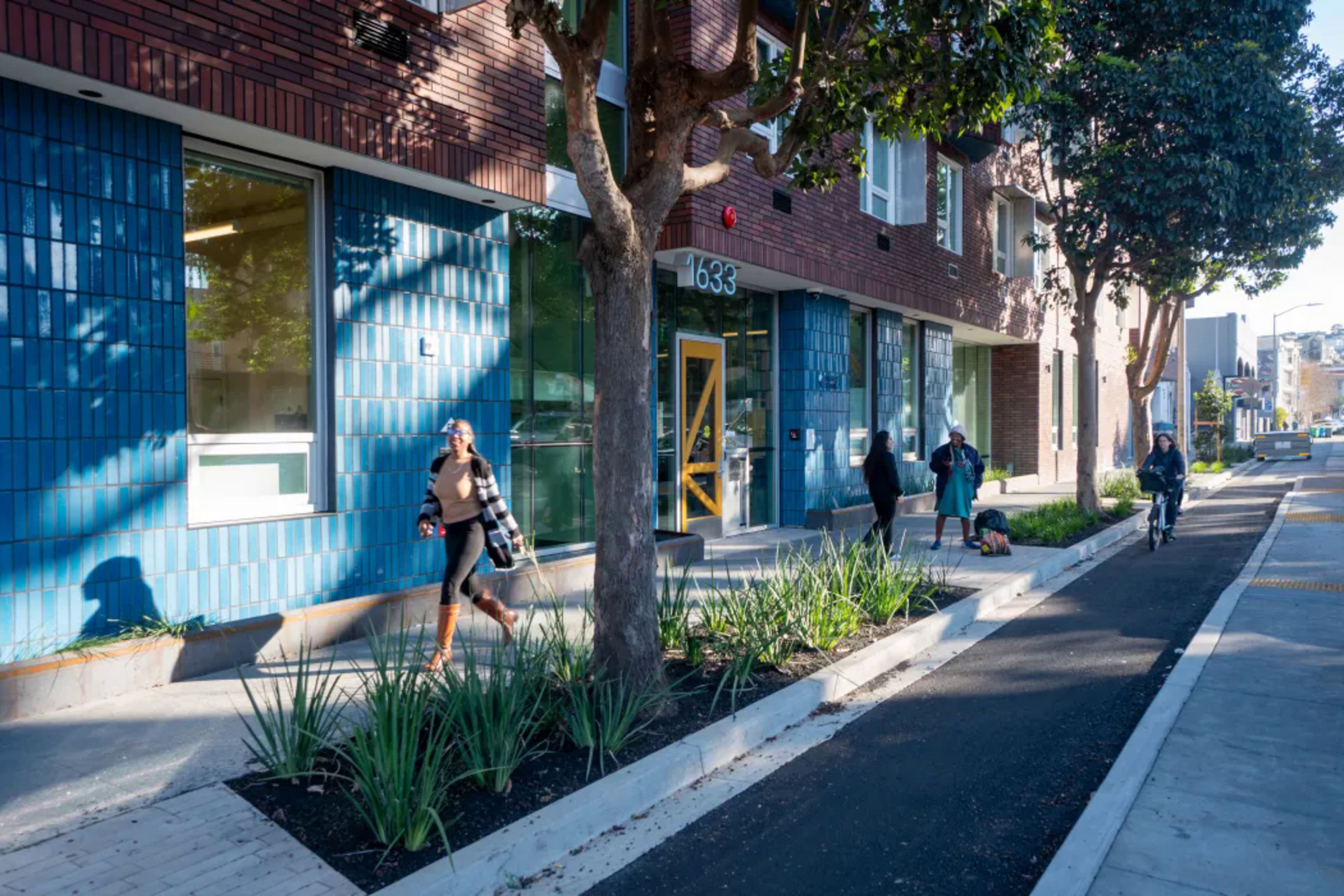 A street scene with a modern brick and blue-tiled building, several trees, plants along the sidewalk, and four people walking or biking.