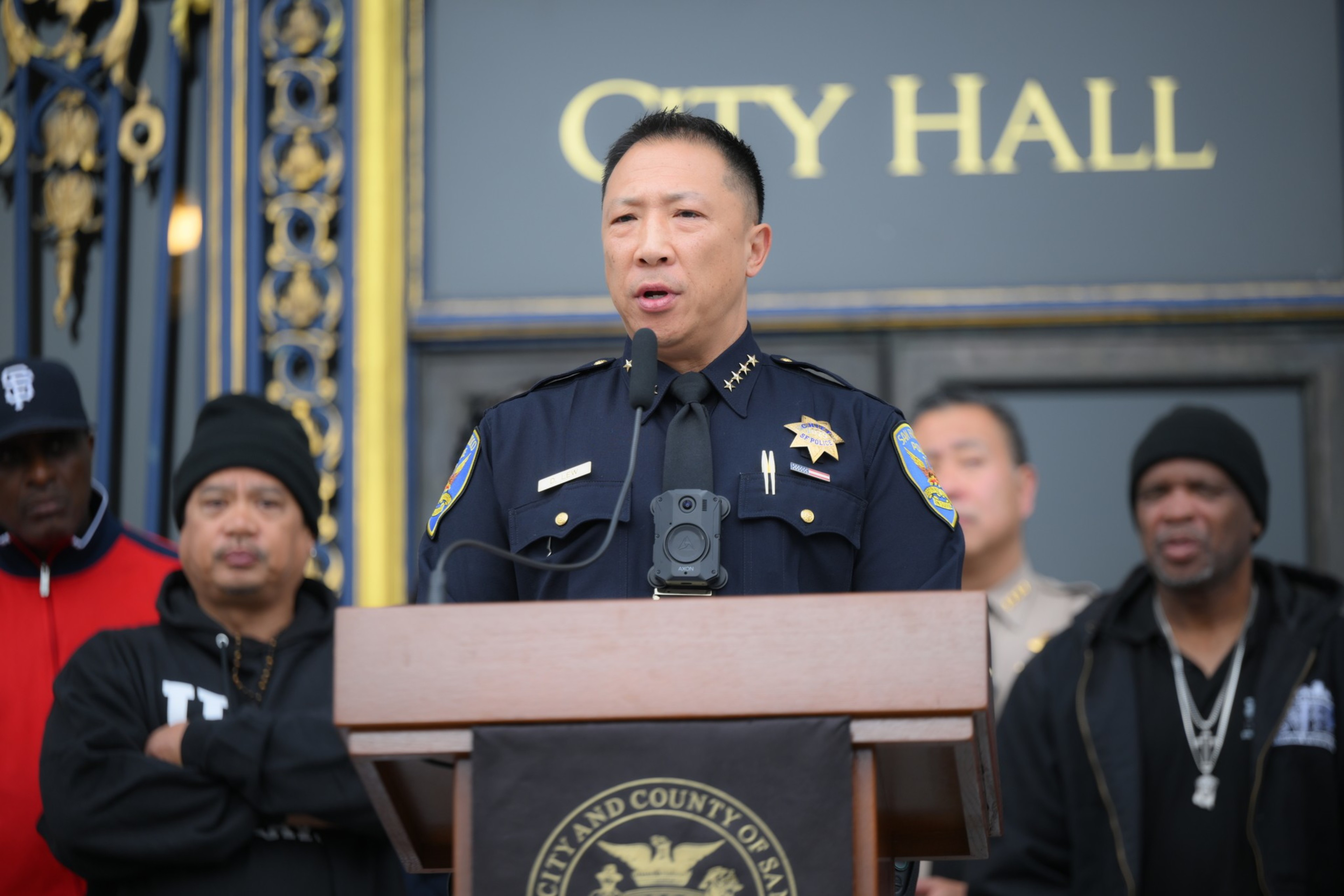 A uniformed police chief speaks at a podium outside City Hall, flanked by men in casual attire and a deputy in uniform.