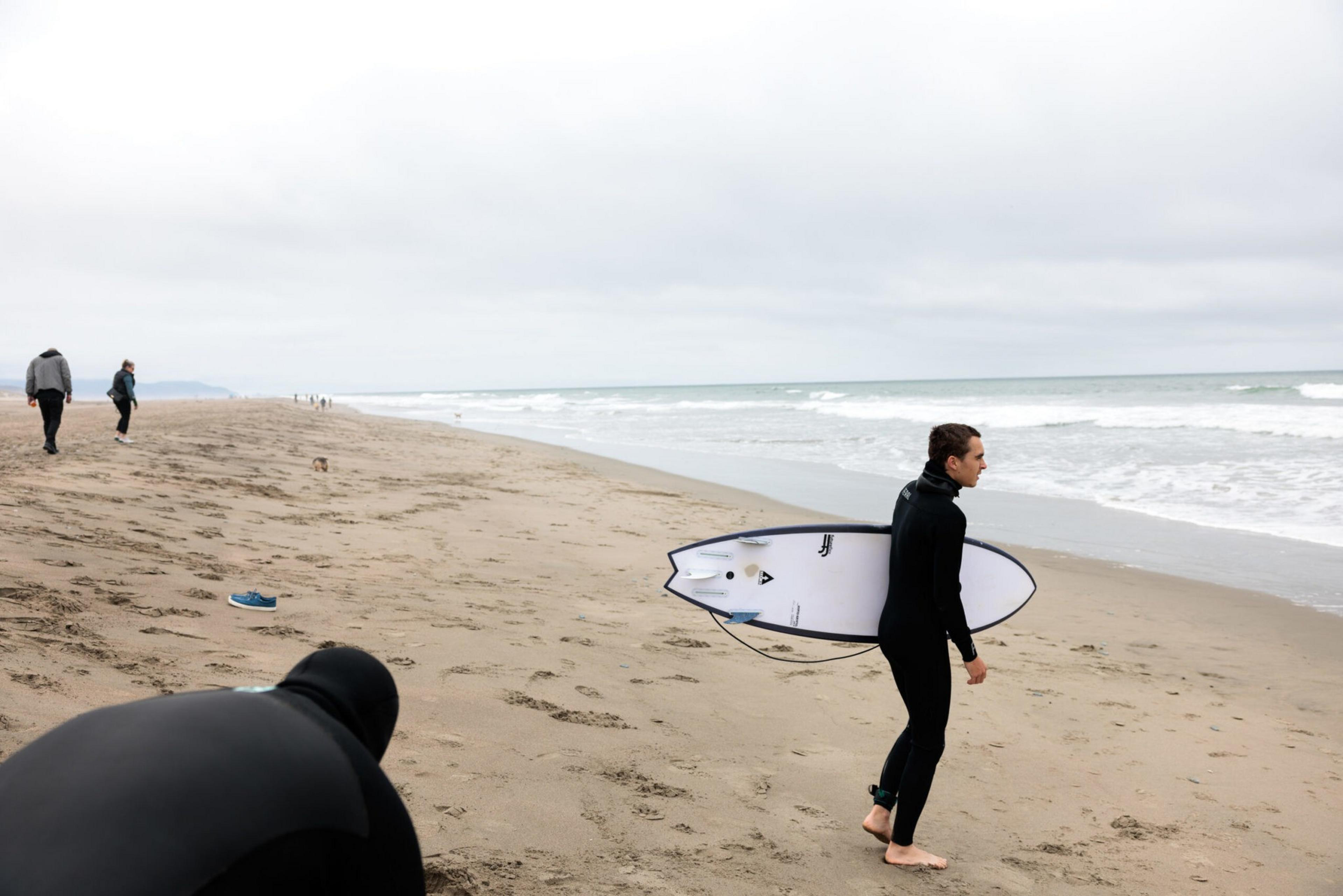 A surfer in a wetsuit carries a white surfboard along a cloudy, sandy beach while other people walk in the distance near the ocean.
