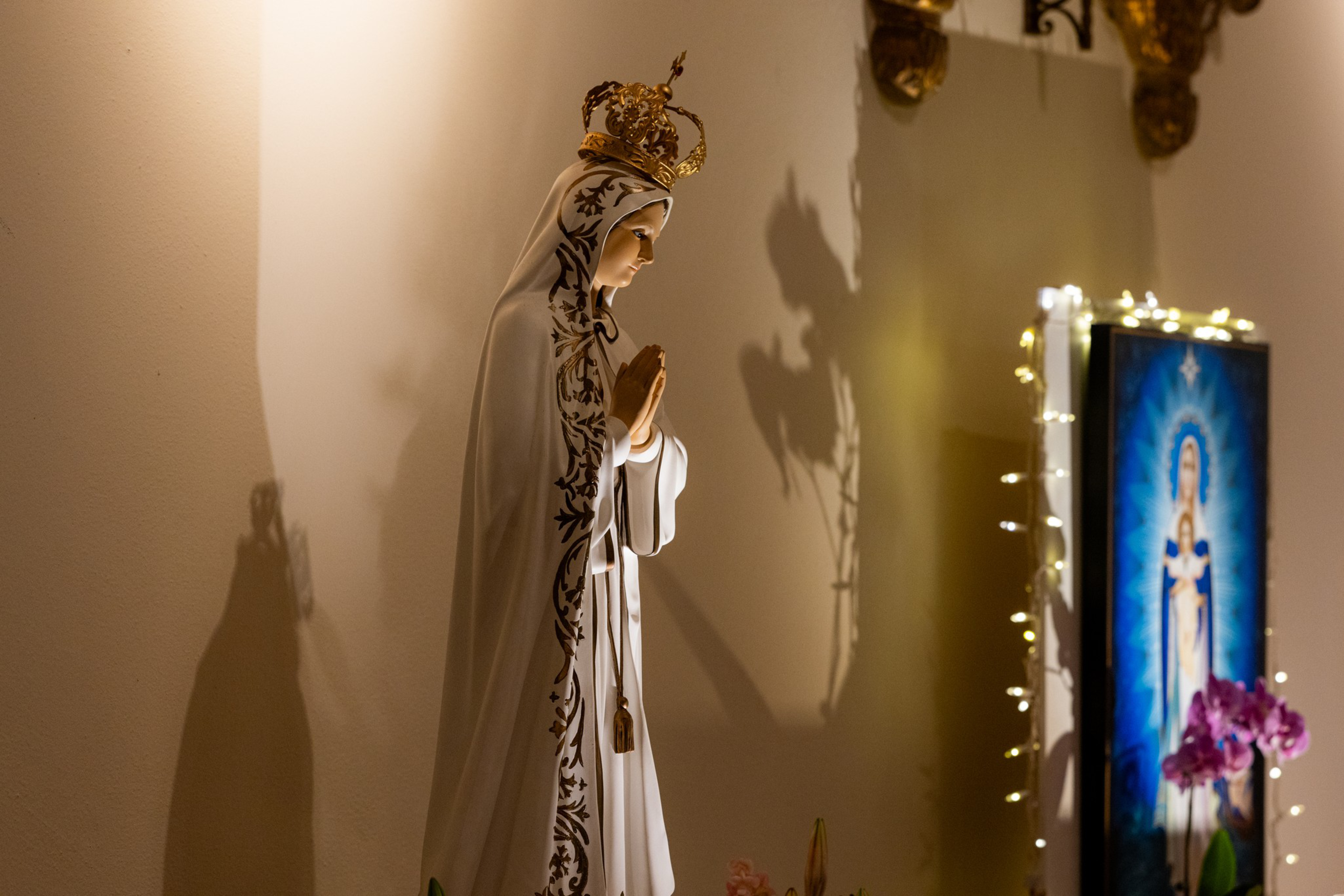 A crowned statue of the Virgin Mary in a white robe with ornate trim stands praying, casting a shadow on the wall near a framed illuminated image and purple flowers.
