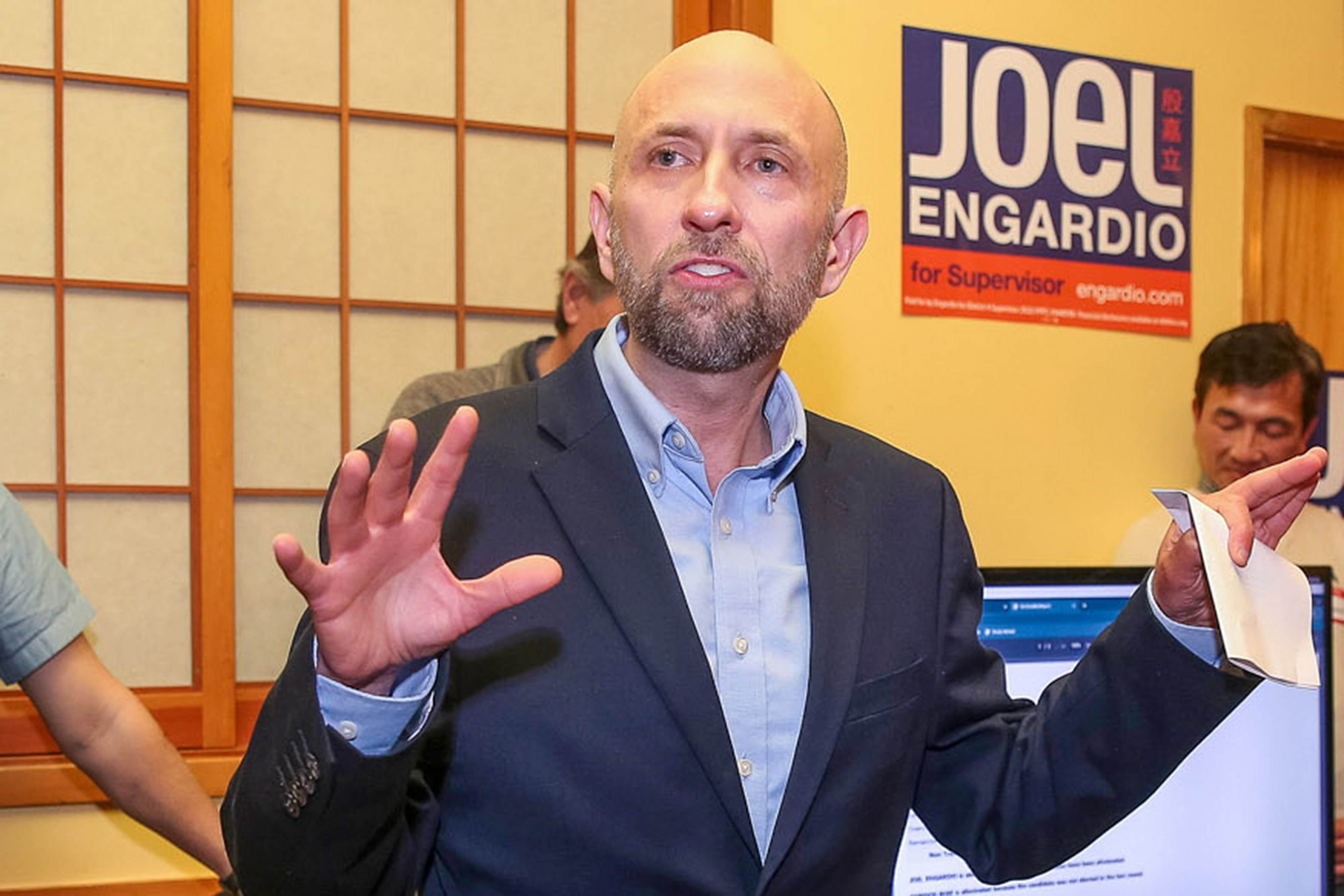 A bald man in a dark blazer and light blue shirt gestures with his hands while speaking in front of a sign reading "Joe Engardio for Supervisor."