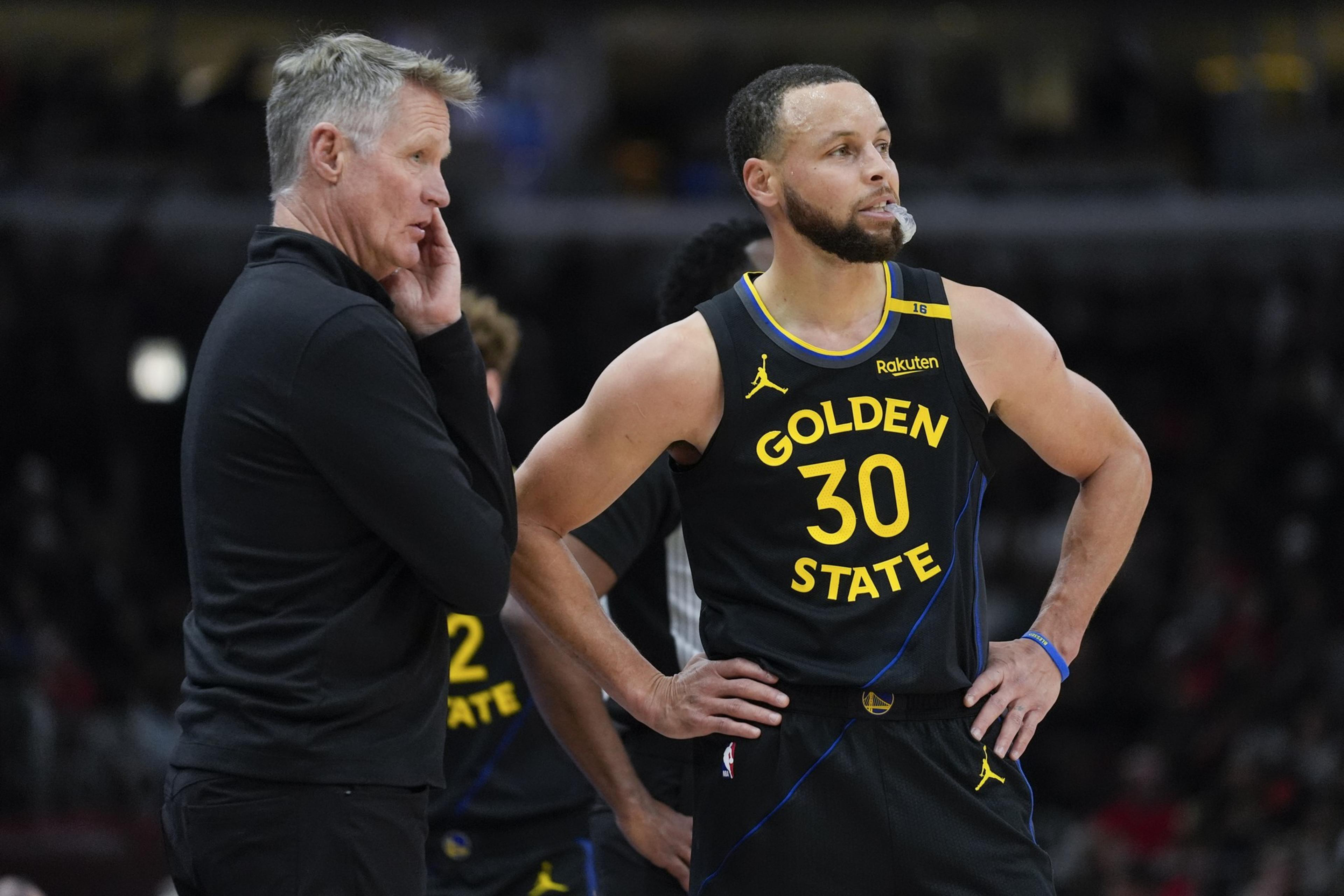 A basketball player in a "Golden State" jersey stands with hands on hips, alongside a man in a black jacket. The focus seems to be on a game or strategy.
