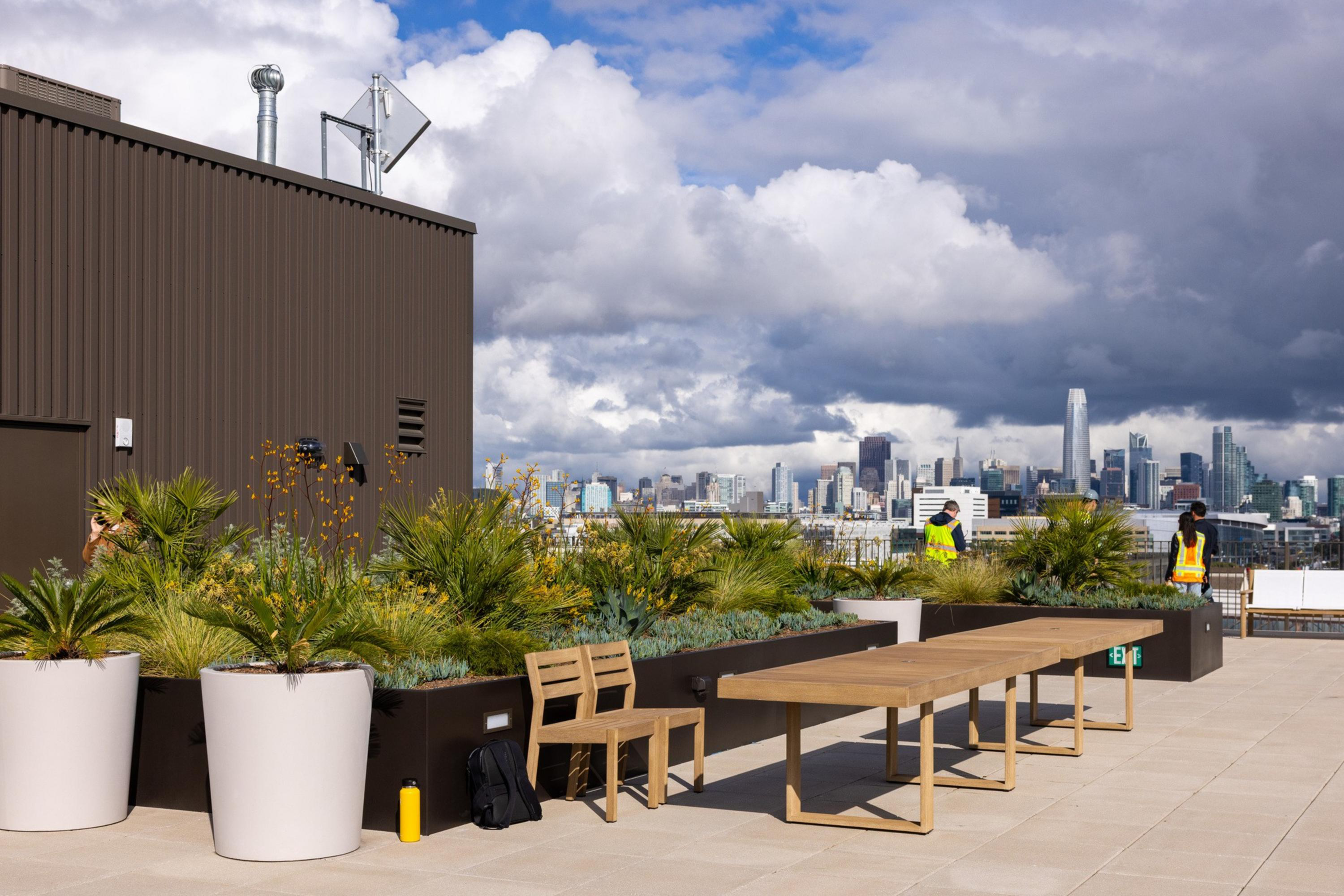 A rooftop patio with wooden chairs, tables, and large planters filled with greenery overlooks a city skyline under dramatic cloudy skies.