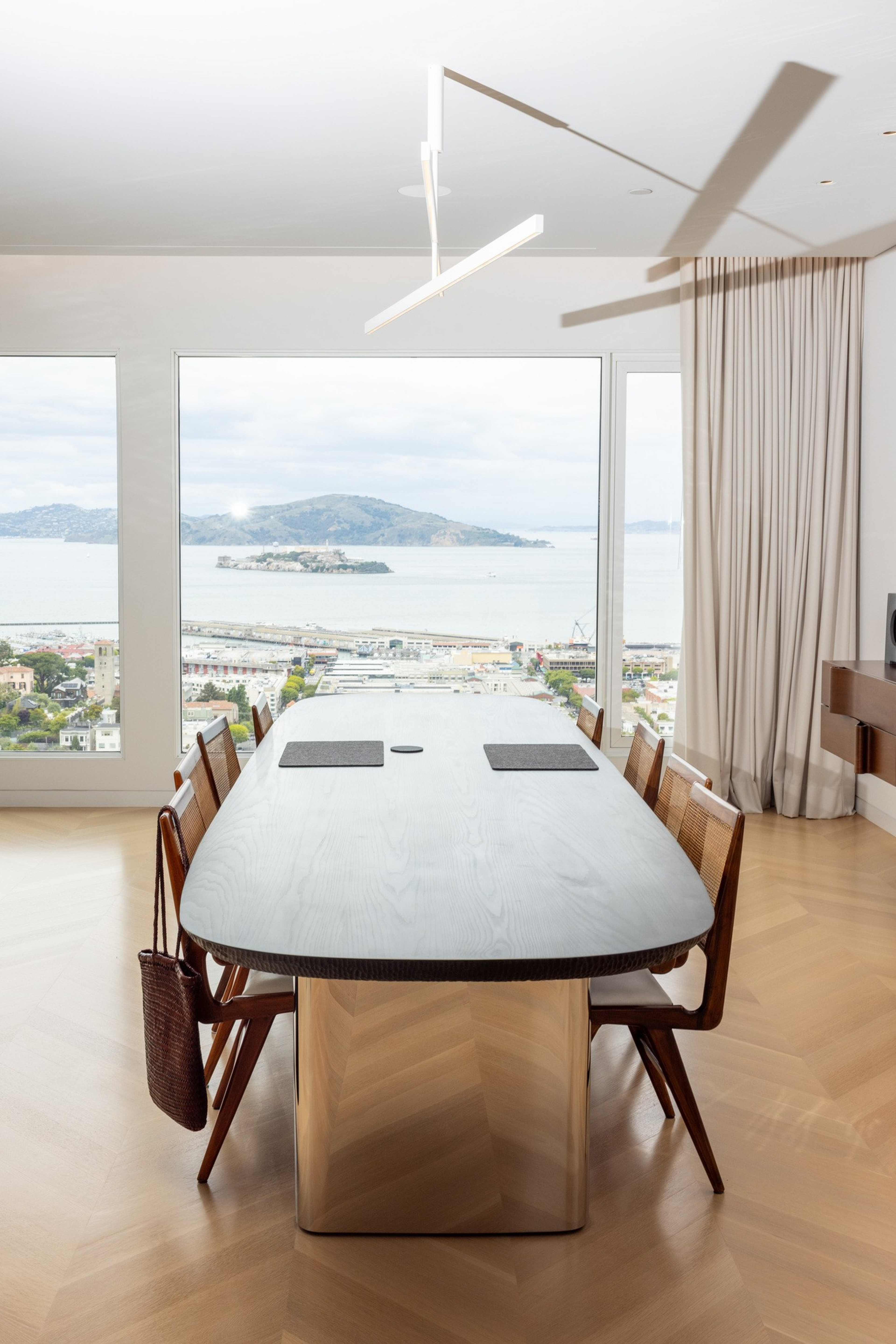 A modern dining table with six wooden chairs faces large windows overlooking a coastal city and hills under a cloudy sky.