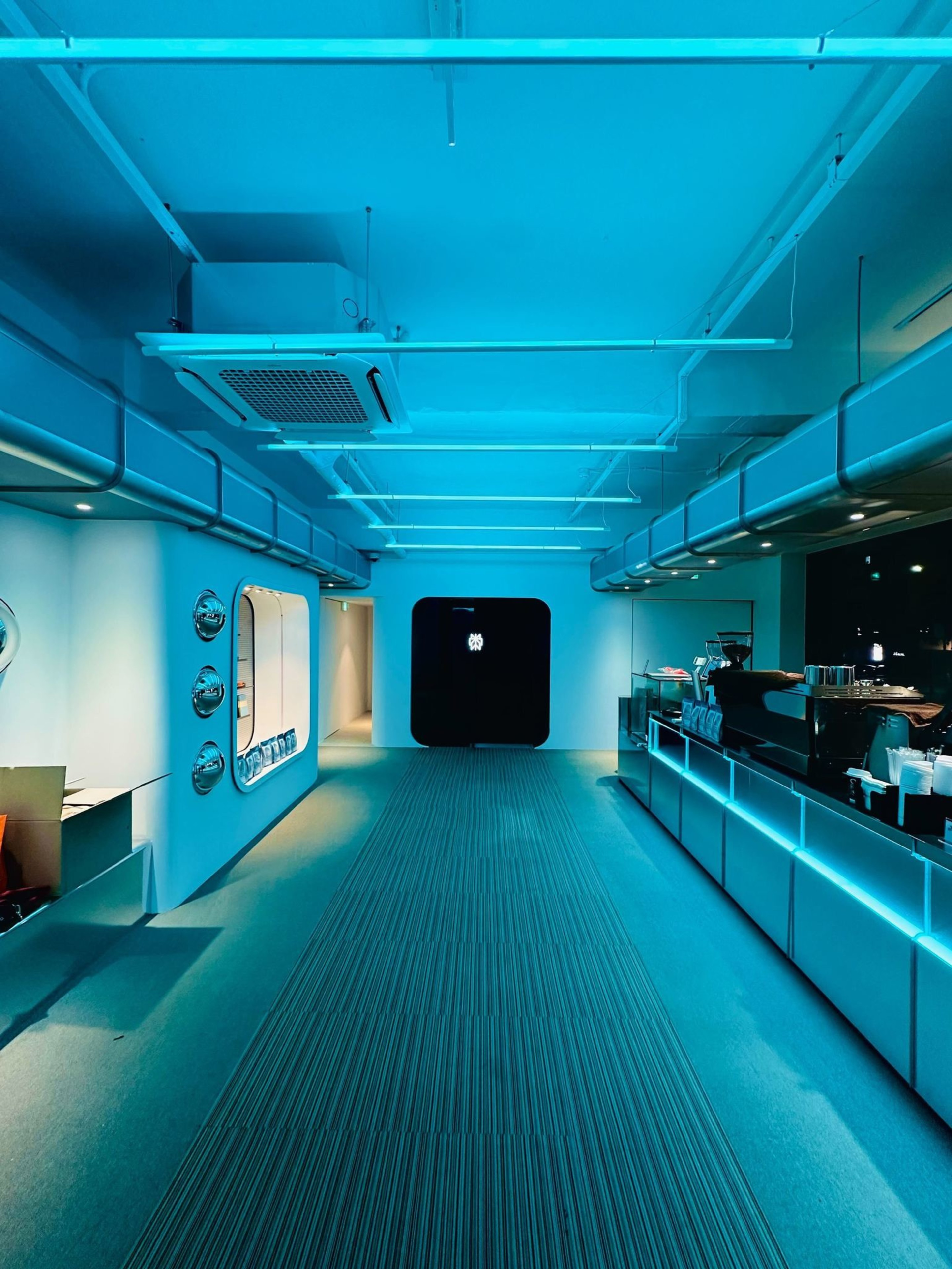 A modern, blue-lit hallway with a carpet runner leads to a large black doorway, with round windows and a sleek counter on the right holding coffee supplies.