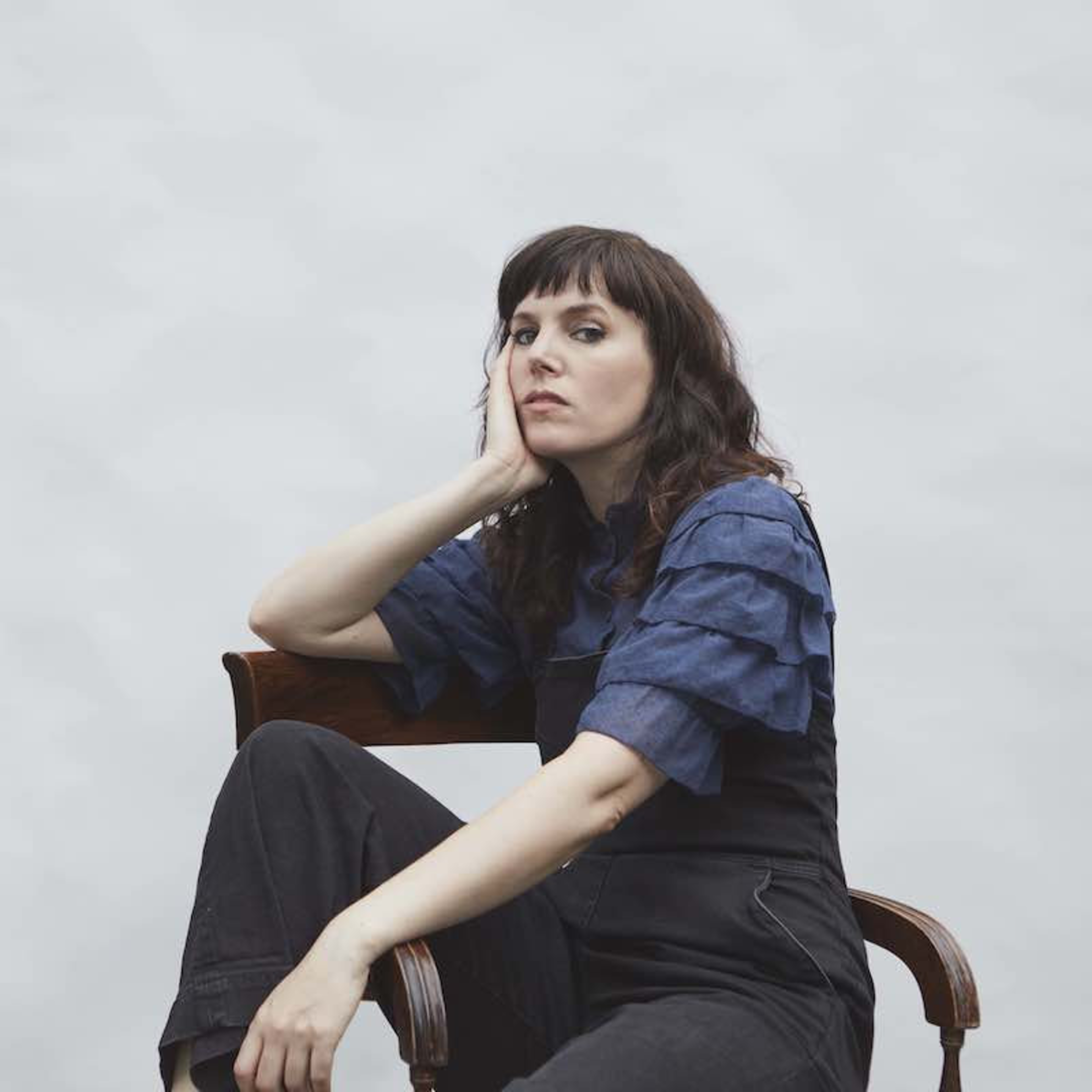 A woman with dark, wavy hair and bangs sits on a wooden chair, resting her head on her hand, wearing a blue ruffled sleeve top and black pants.