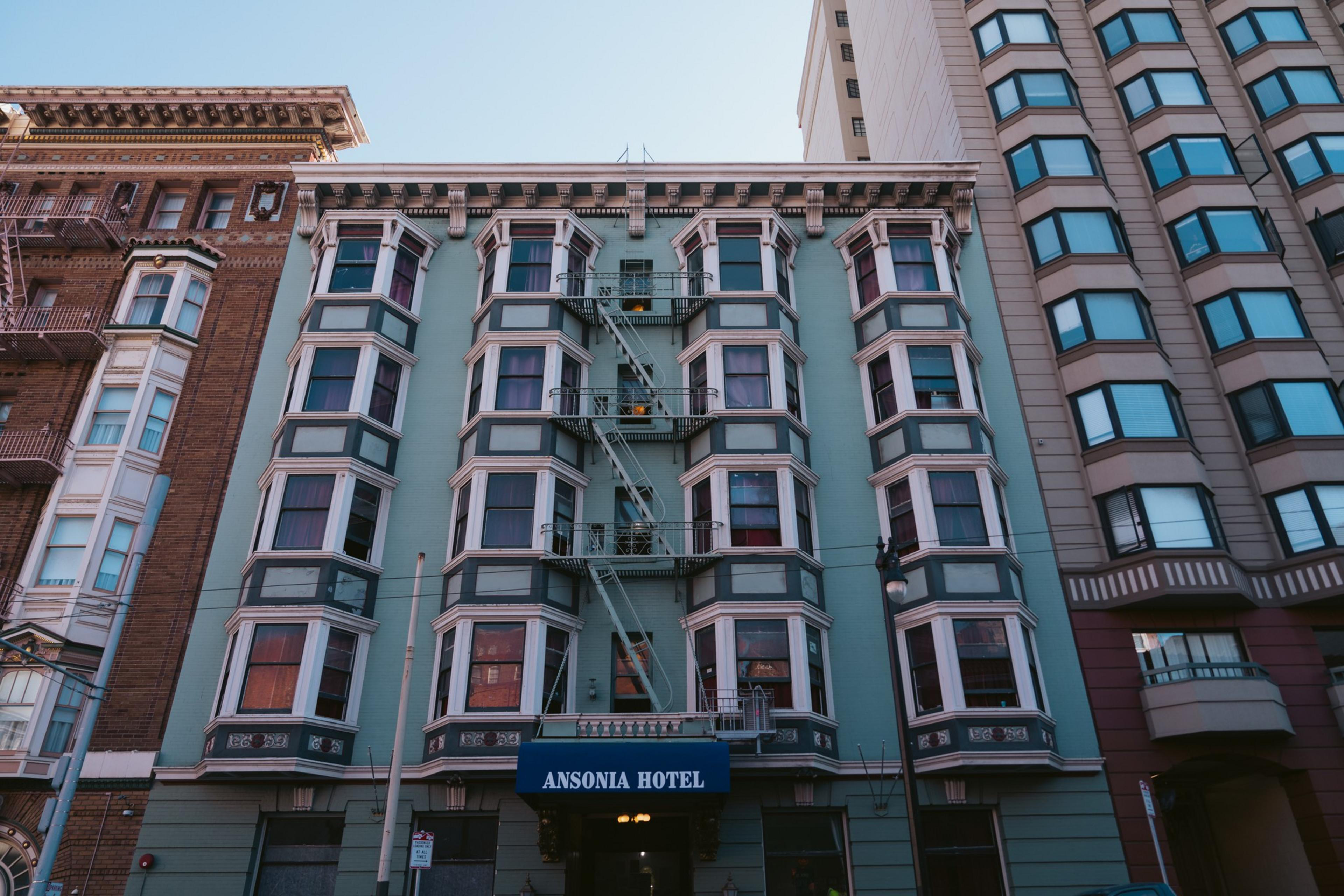 A blue building with white-trimmed bay windows, fire escapes, and a sign reading "Ansonia Hotel" stands between taller modern and classic buildings.