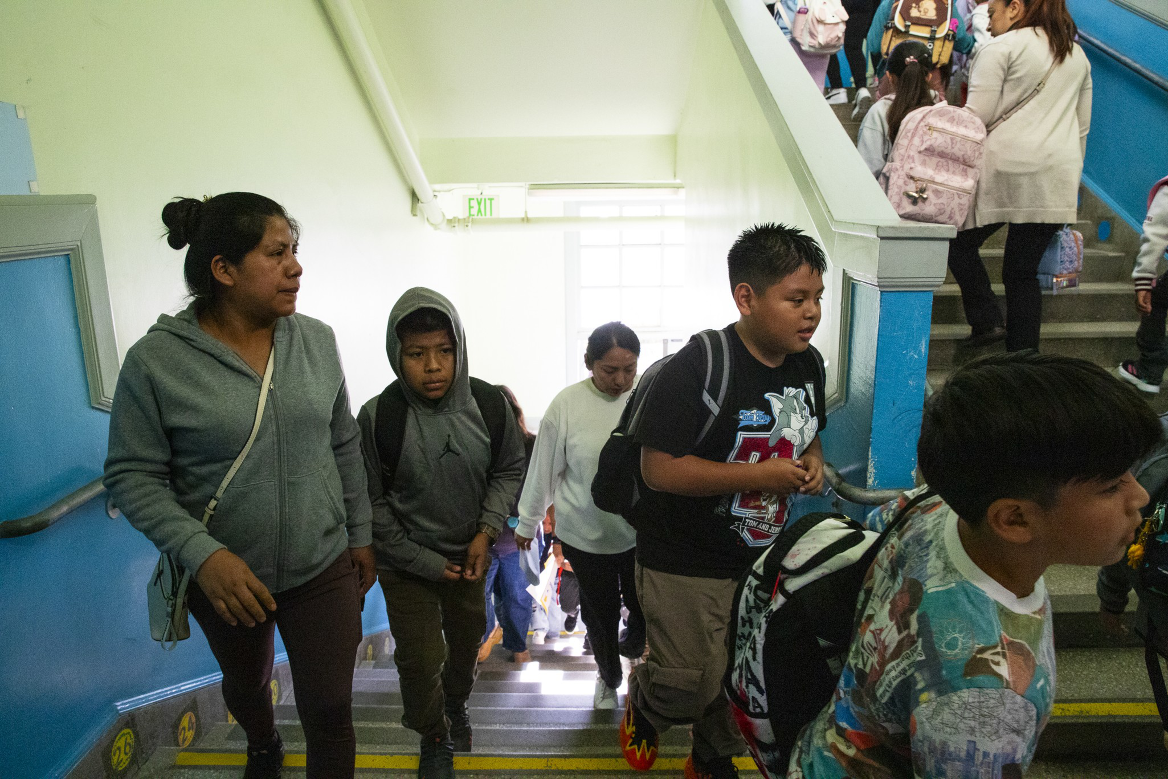 A group of children and adults walk up a staircase inside a brightly lit building, some carrying backpacks.