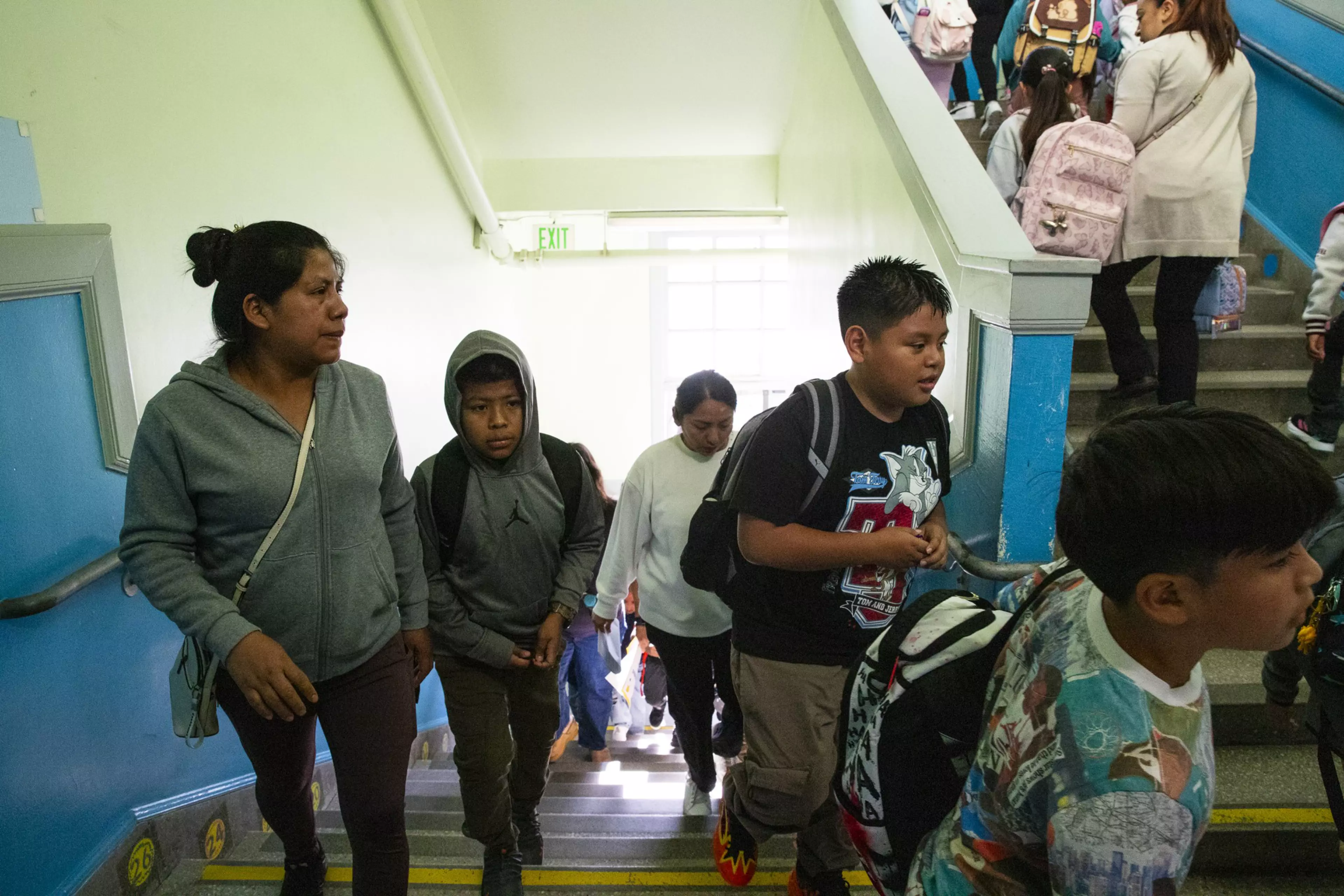 A group of children and adults walk up a staircase inside a brightly lit building, some carrying backpacks.