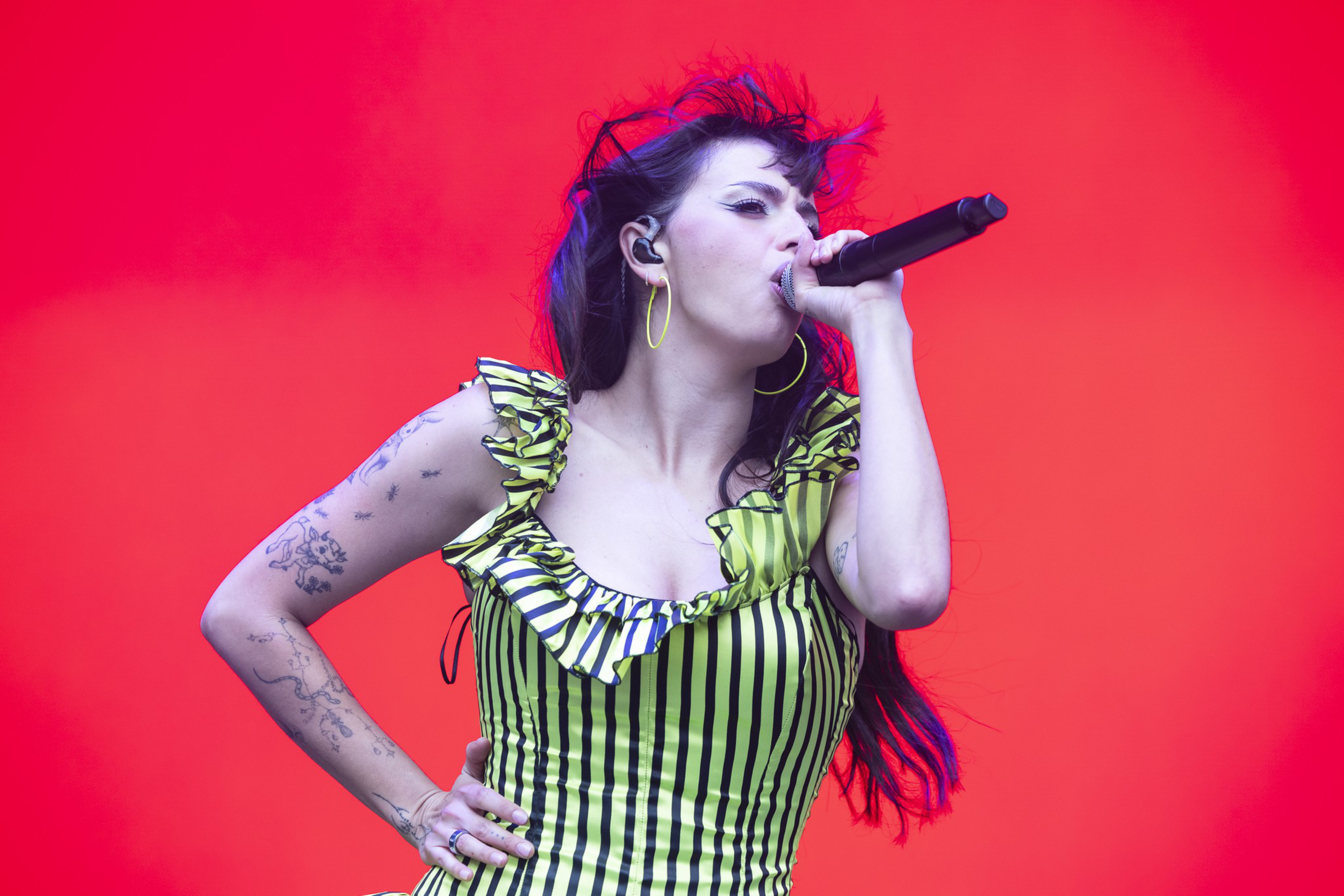 A woman with long dark hair sings into a microphone against a bright red background, wearing a green and black striped ruffled top and hoop earrings.