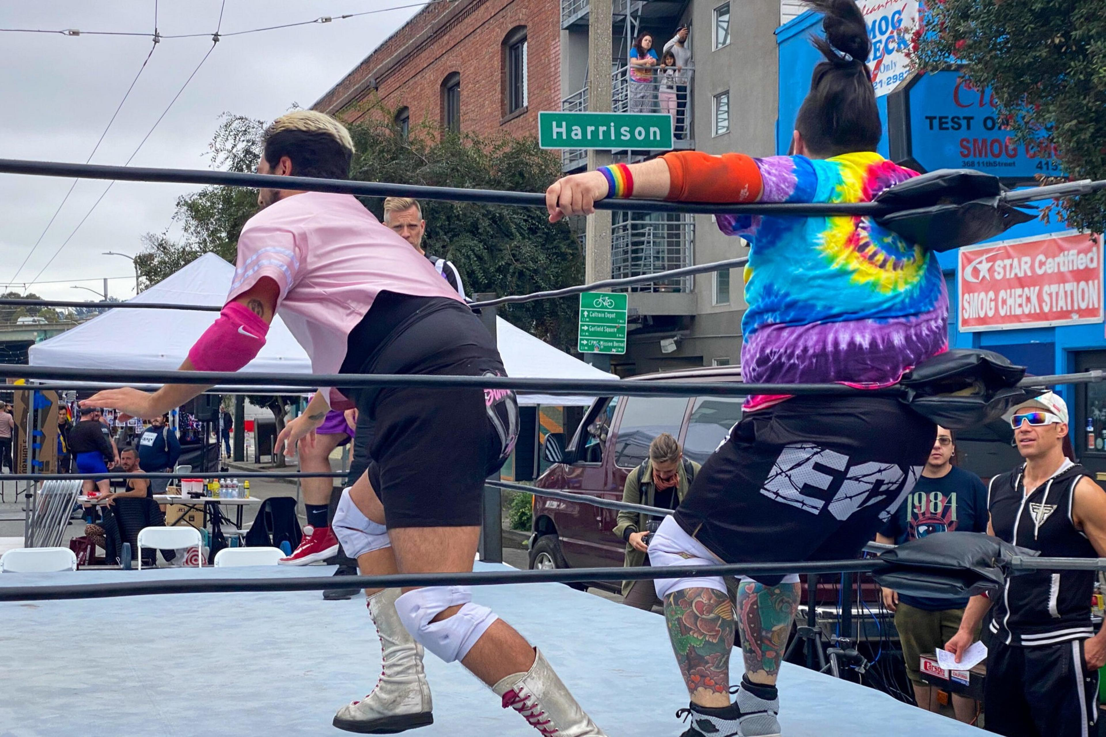 Two wrestlers, one in a tie-dye shirt and the other in pink, face off inside a wrestling ring during an outdoor event on Harrison street.
