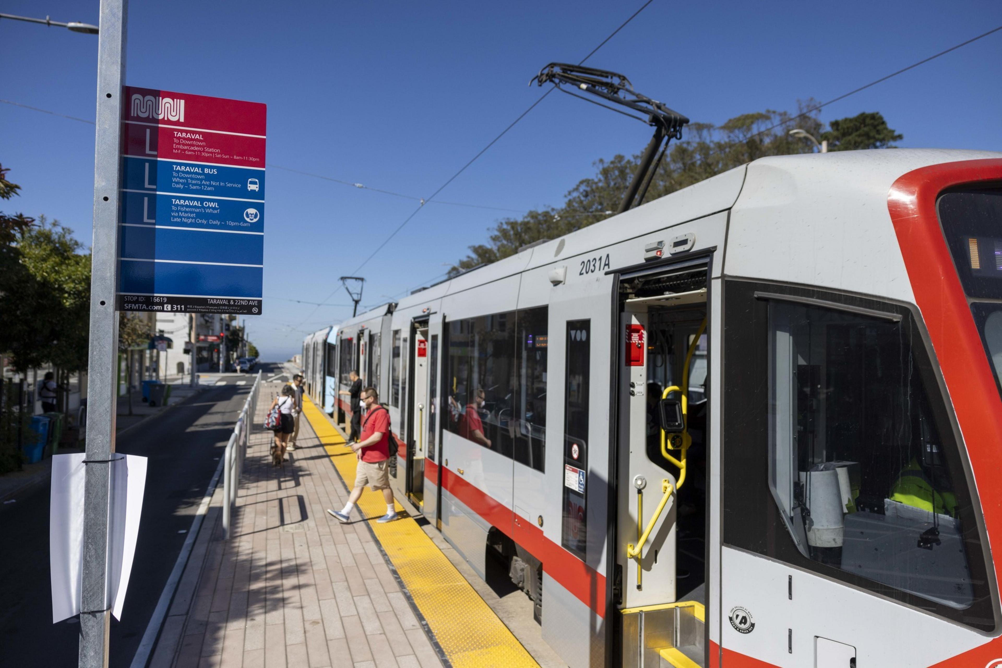 A white and red train is stopped at a station, with its doors open. Several passengers are boarding. A nearby pole displays a "Muni" sign with route information.