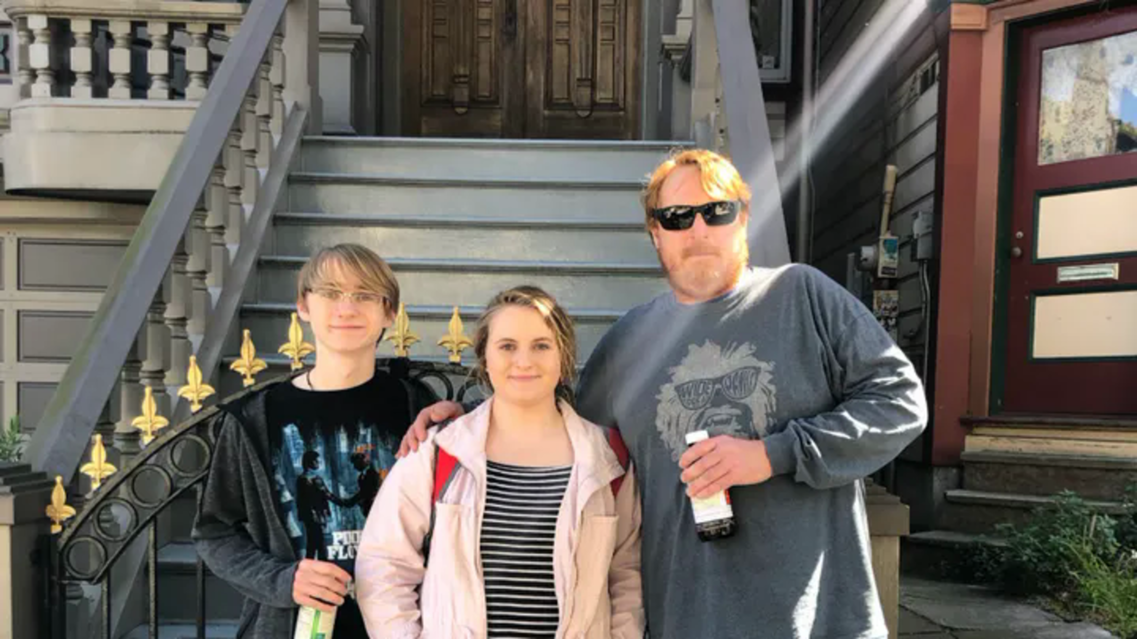 Three people stand closely together in front of a house with stone steps, wearing casual clothes, with the two men holding bottled drinks.