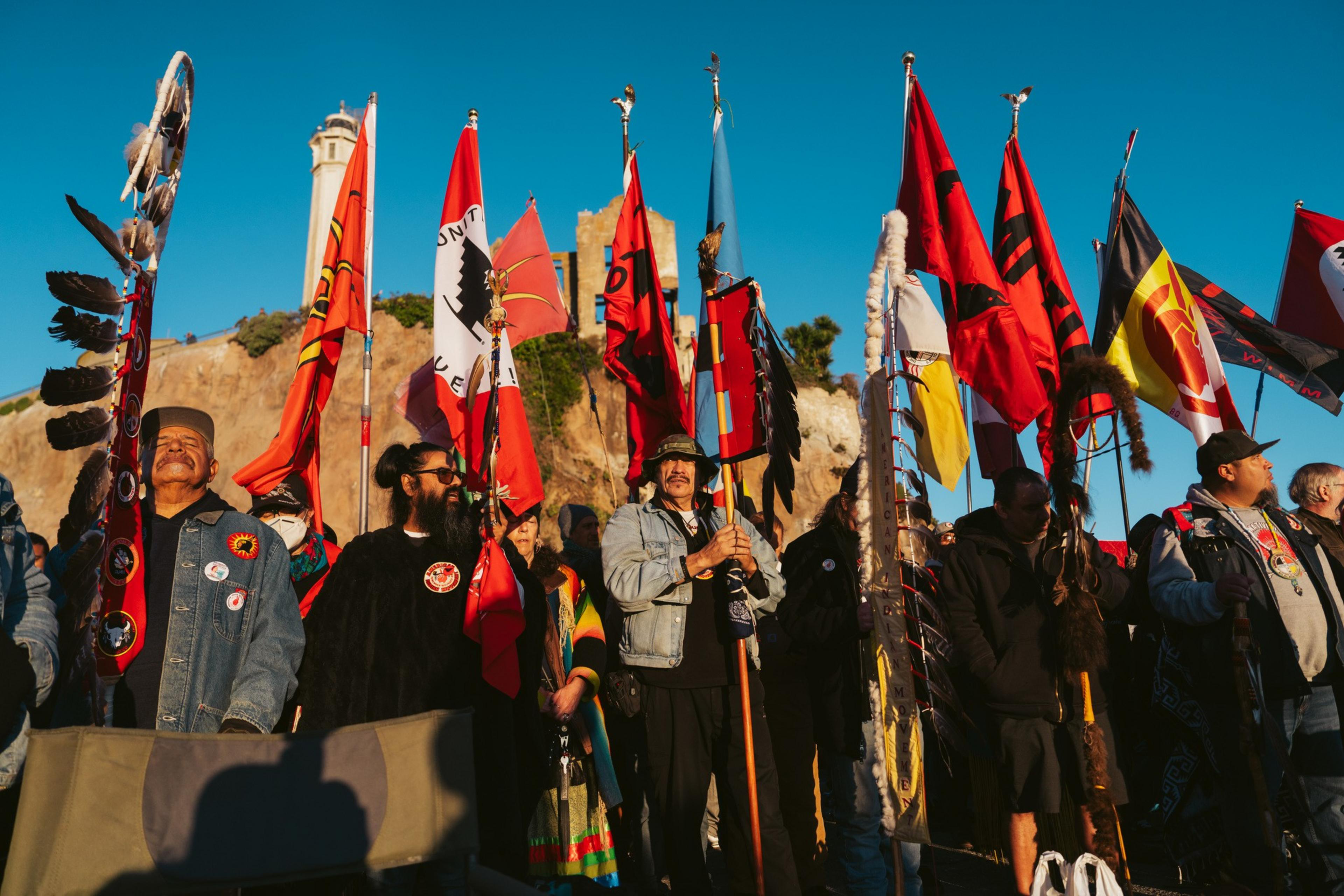 A group of people stands outdoors holding vibrant flags, against a backdrop of a rocky hill and a blue sky. Some flags are adorned with feathers.