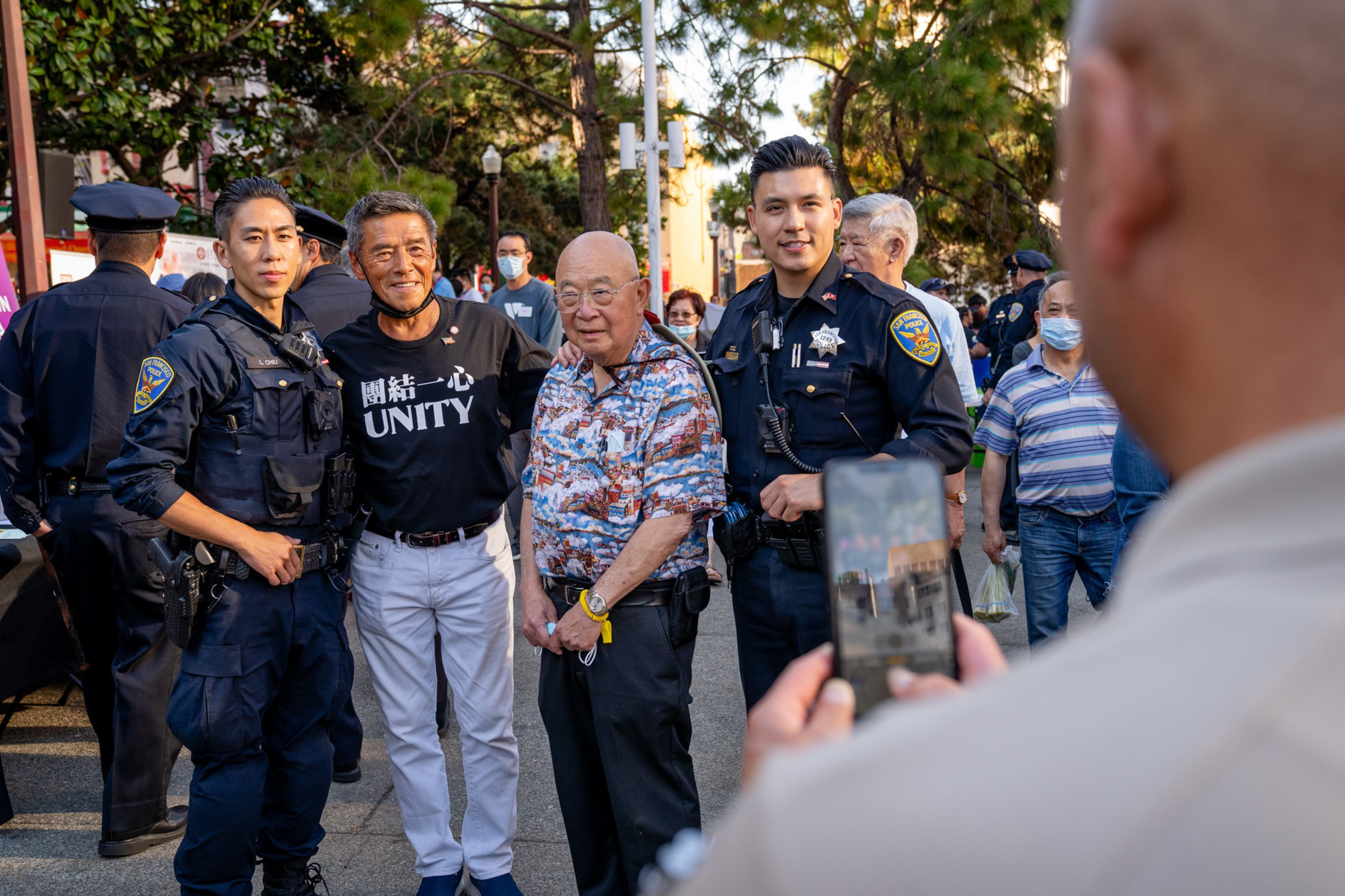 Three men pose with two police officers while another person takes their photo using a smartphone in an outdoor, crowded setting.
