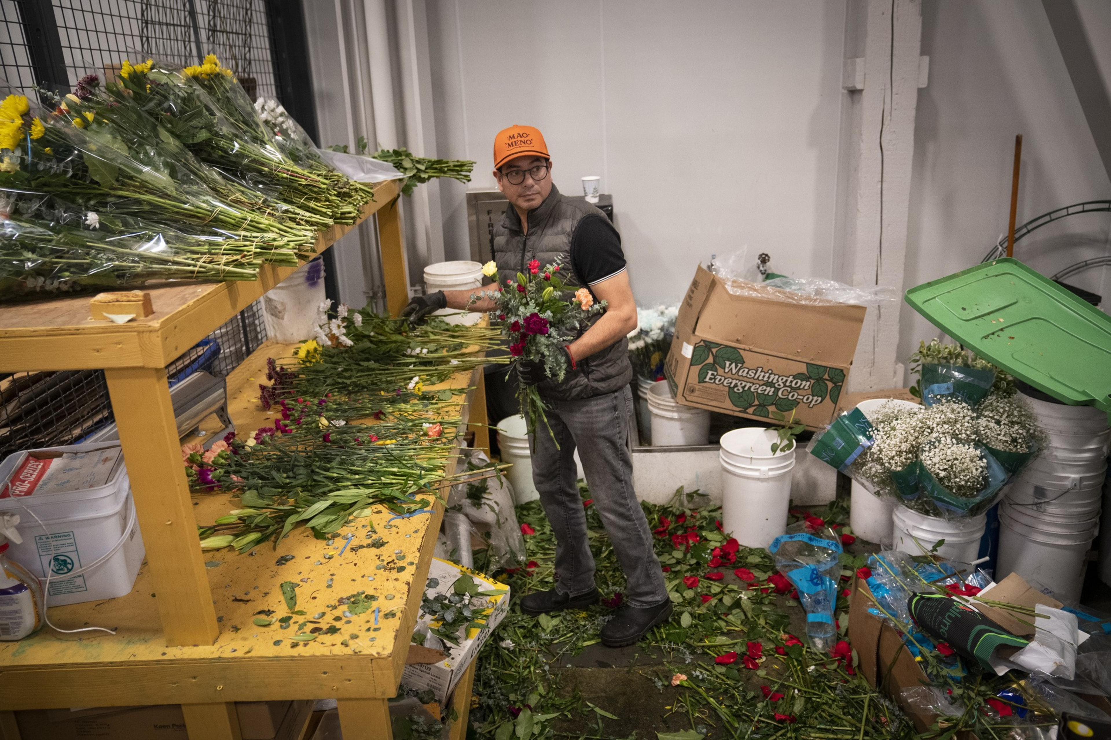 A person stands in a workshop arranging flowers, surrounded by shelves of floral bundles and scattered petals. They wear an orange cap and a dark vest.