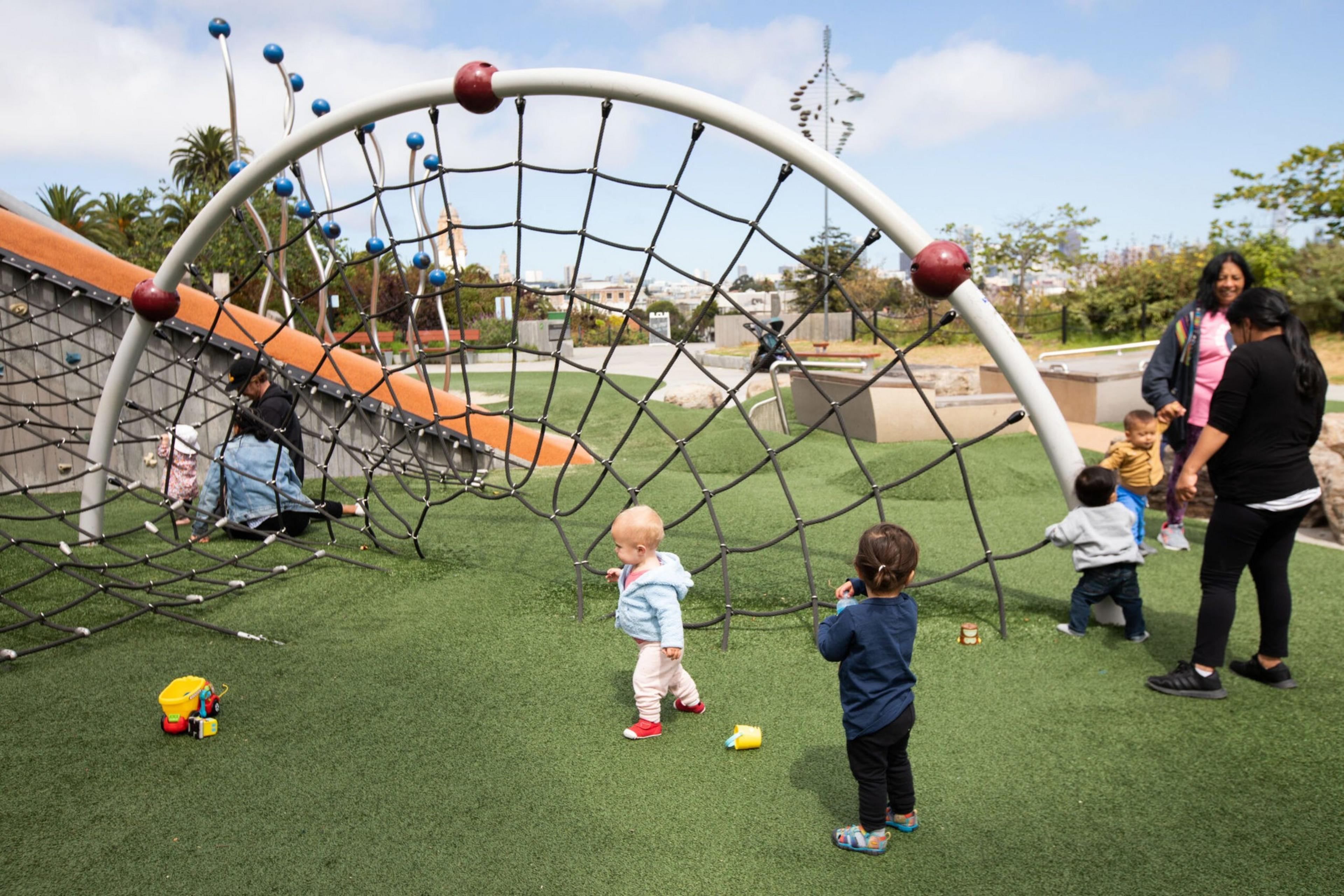 Several toddlers and two adults are playing near a spider-web style climbing structure on a playground with artificial grass.