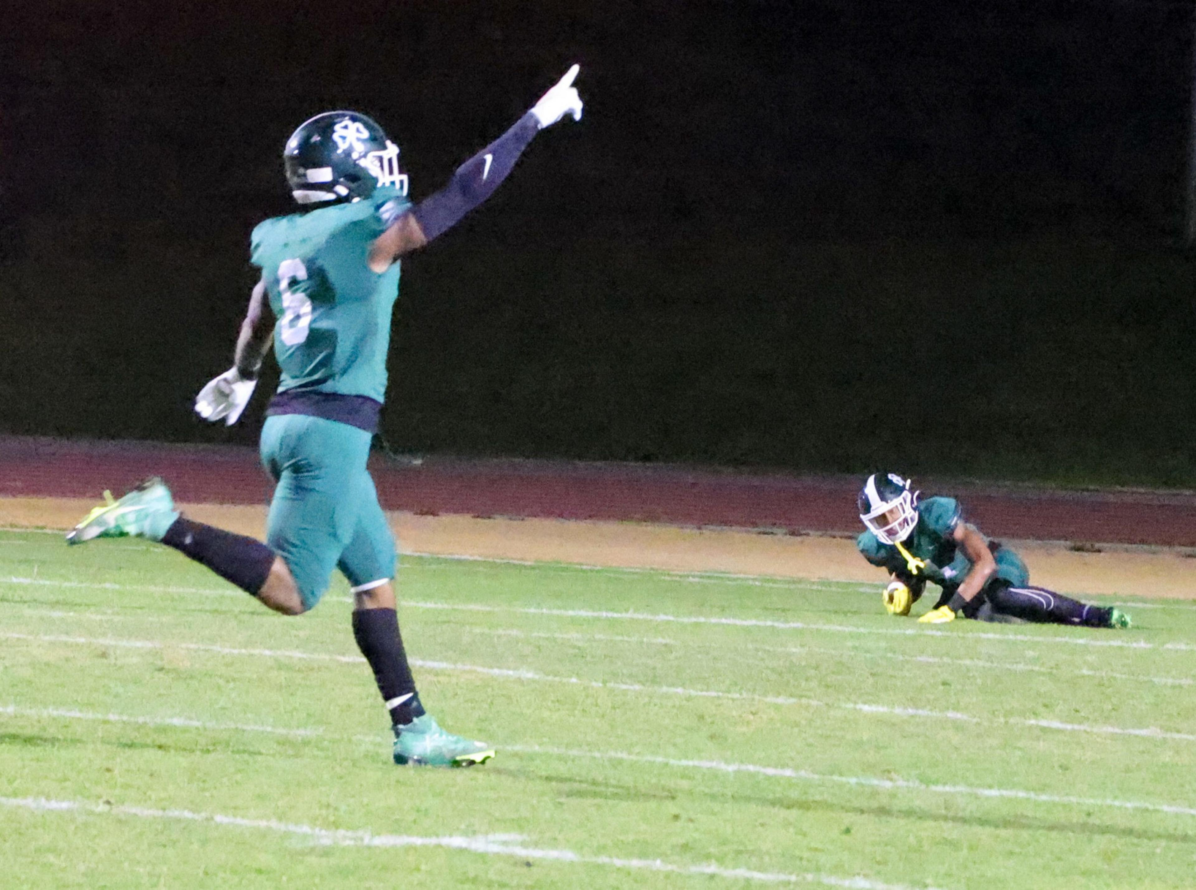 A football player wearing number 6 runs celebrating while a teammate kneels on the ground, holding a football securely.