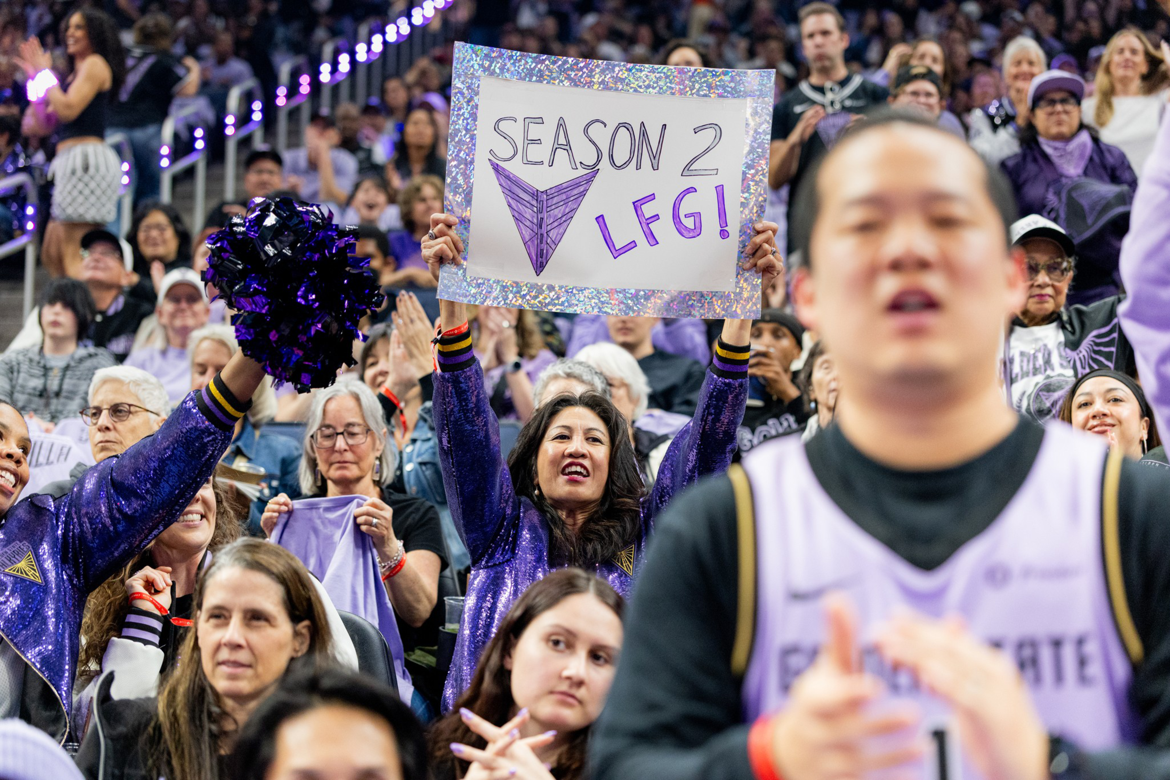 A cheering crowd at a basketball game includes a woman holding a colorful sign that says “Season 2 LEG!” while fans around her wear purple.