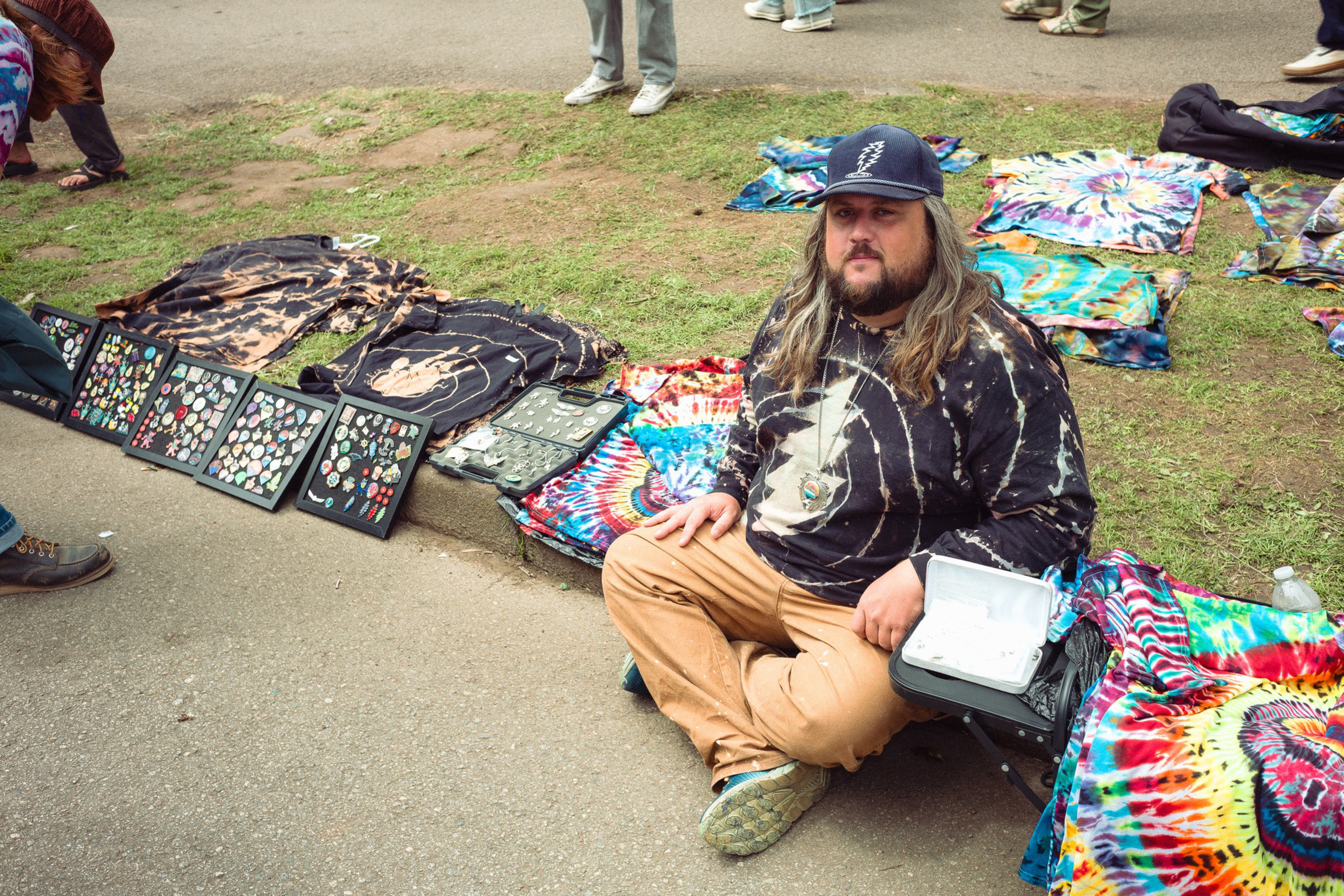 A man in a tie-dye shirt and cap sits on the ground selling colorful tie-dye clothes and framed pin collections at an outdoor market.