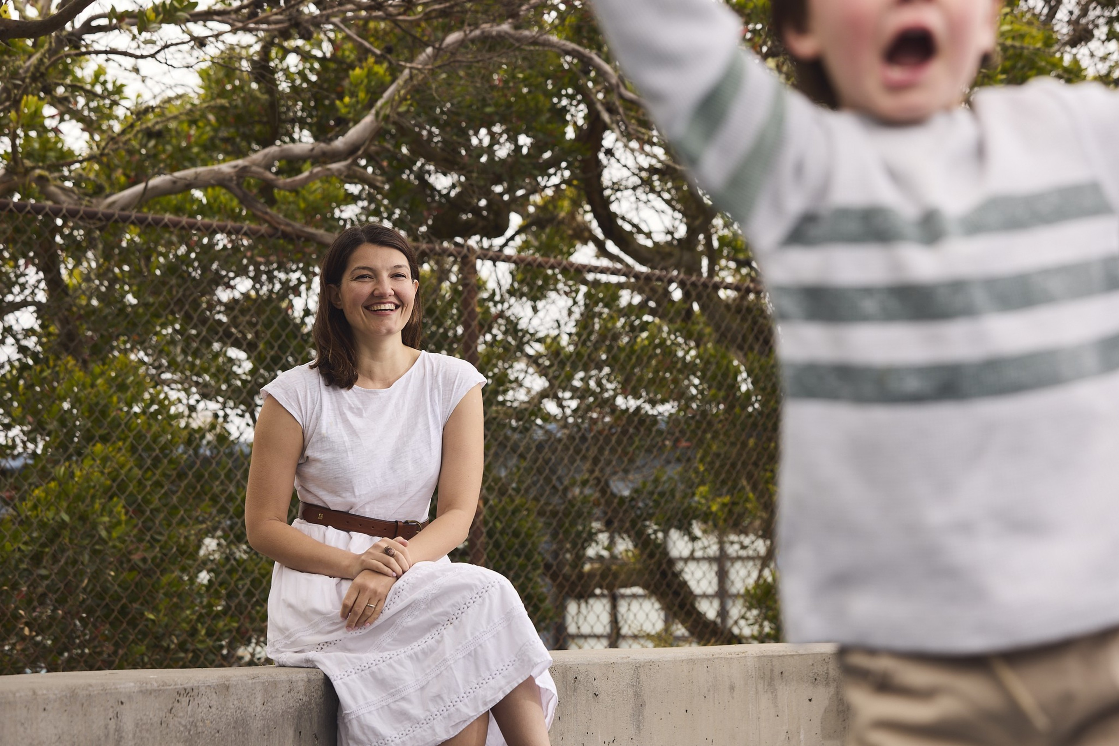 A woman in a white dress sits smiling on a concrete ledge with trees and a wire fence behind her, while a blurred child in a striped sweater moves in the foreground.