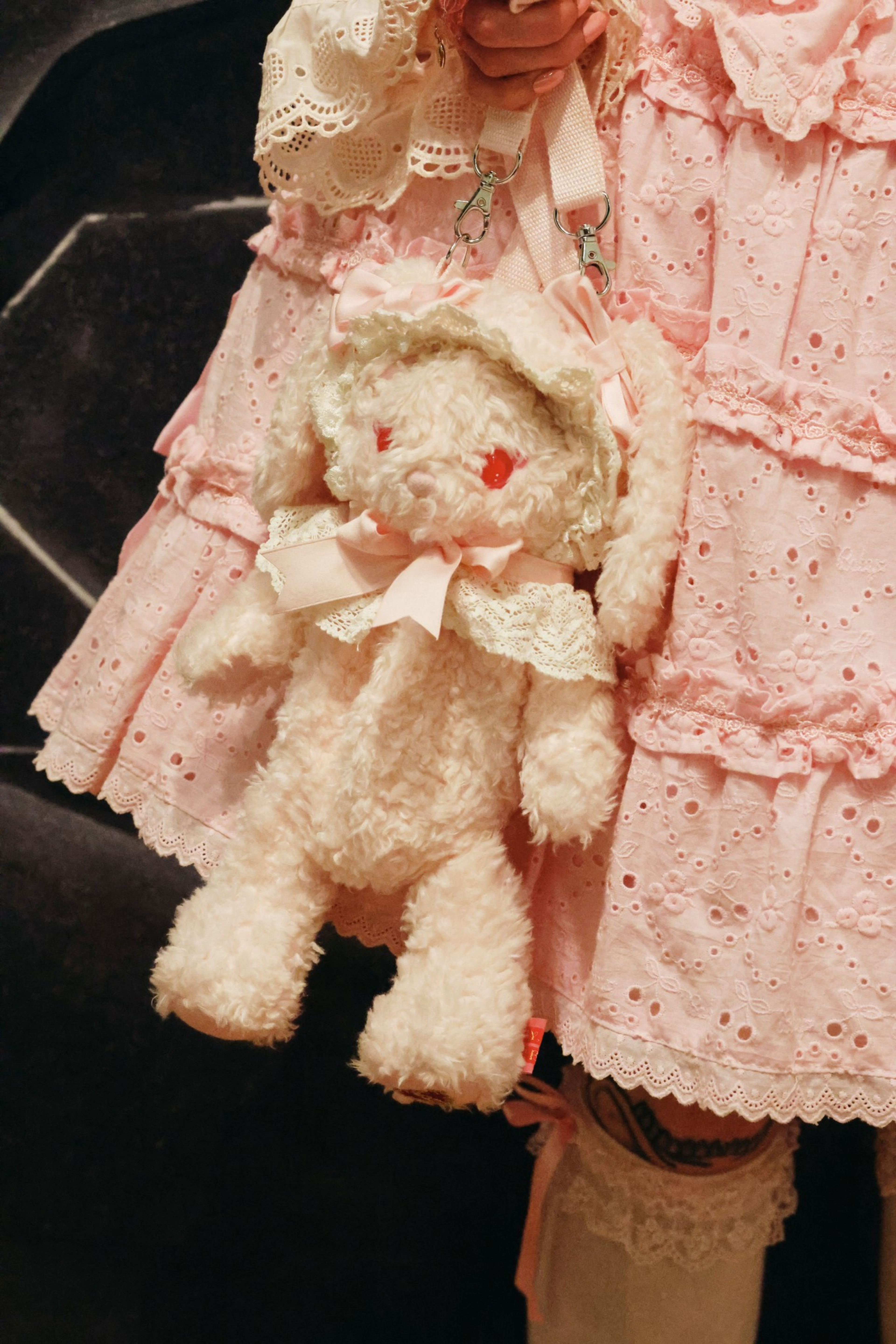 A fluffy teddy bear bag with red eyes and a lace bonnet hangs from a person wearing a pink, eyelet lace dress with ruffles.
