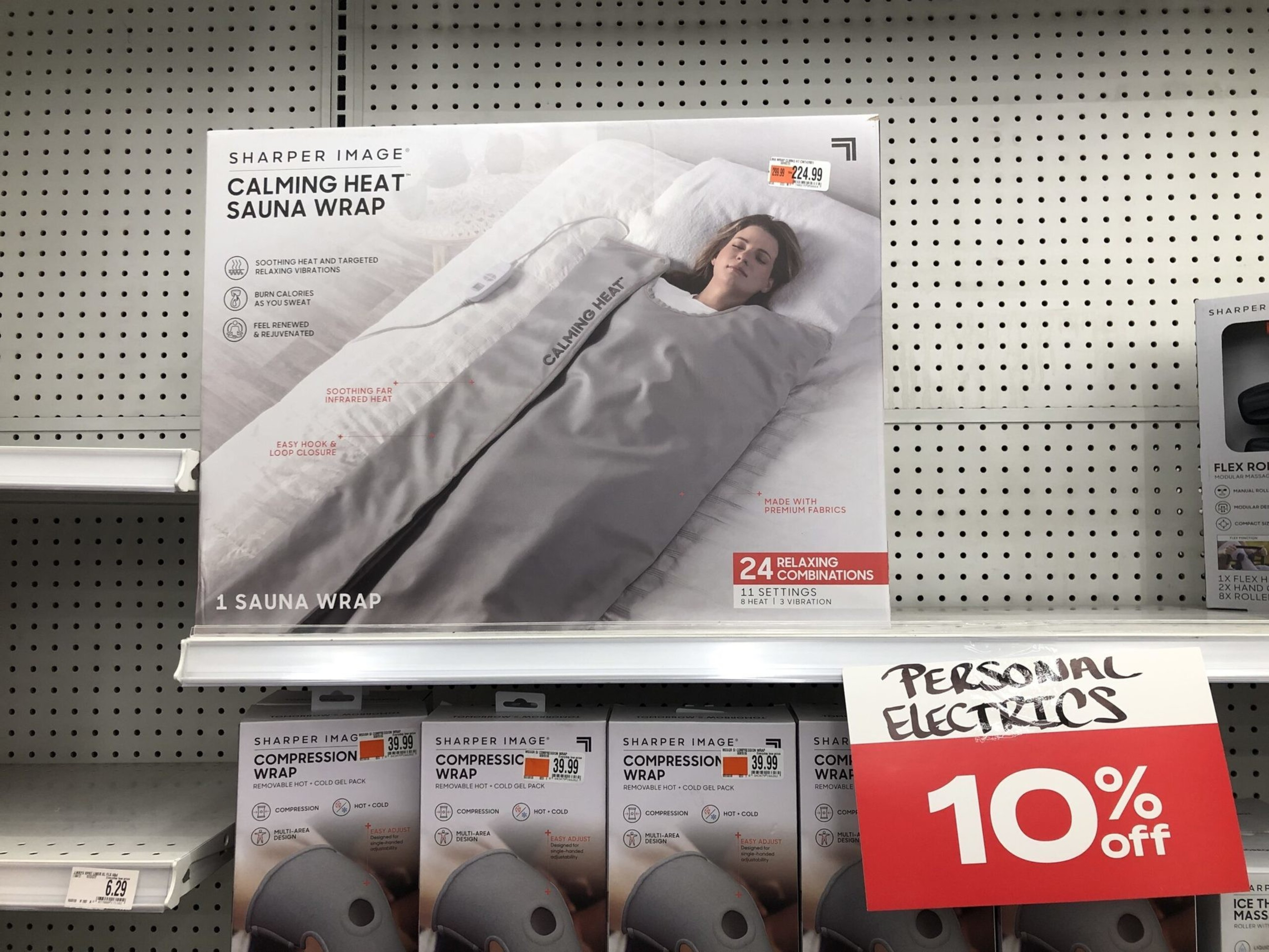 A Sharper Image Calming Heat Sauna Wrap box with a woman wrapped in a gray heated wrap is displayed on a shelf with a 10% off Personal Electrics sign.