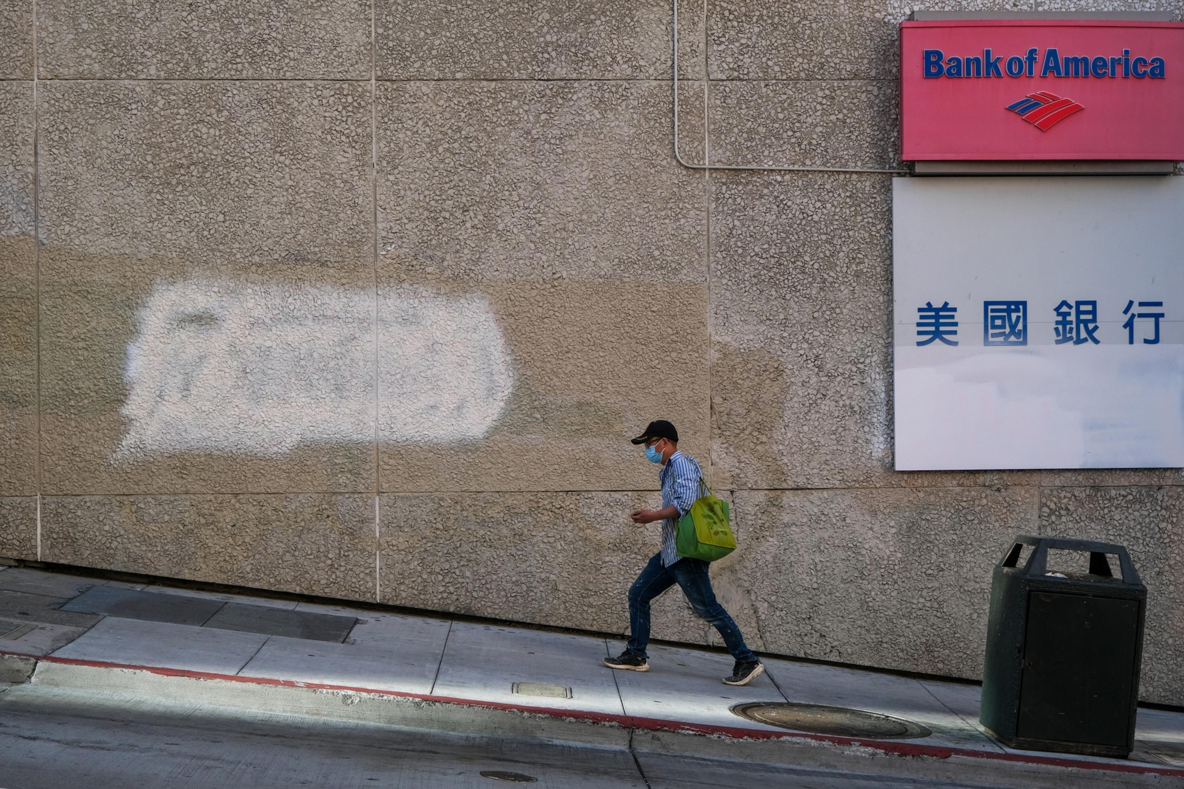 A person wearing a mask, hat, and carrying a green bag walks on a sloped sidewalk next to a Bank of America sign on a concrete wall.