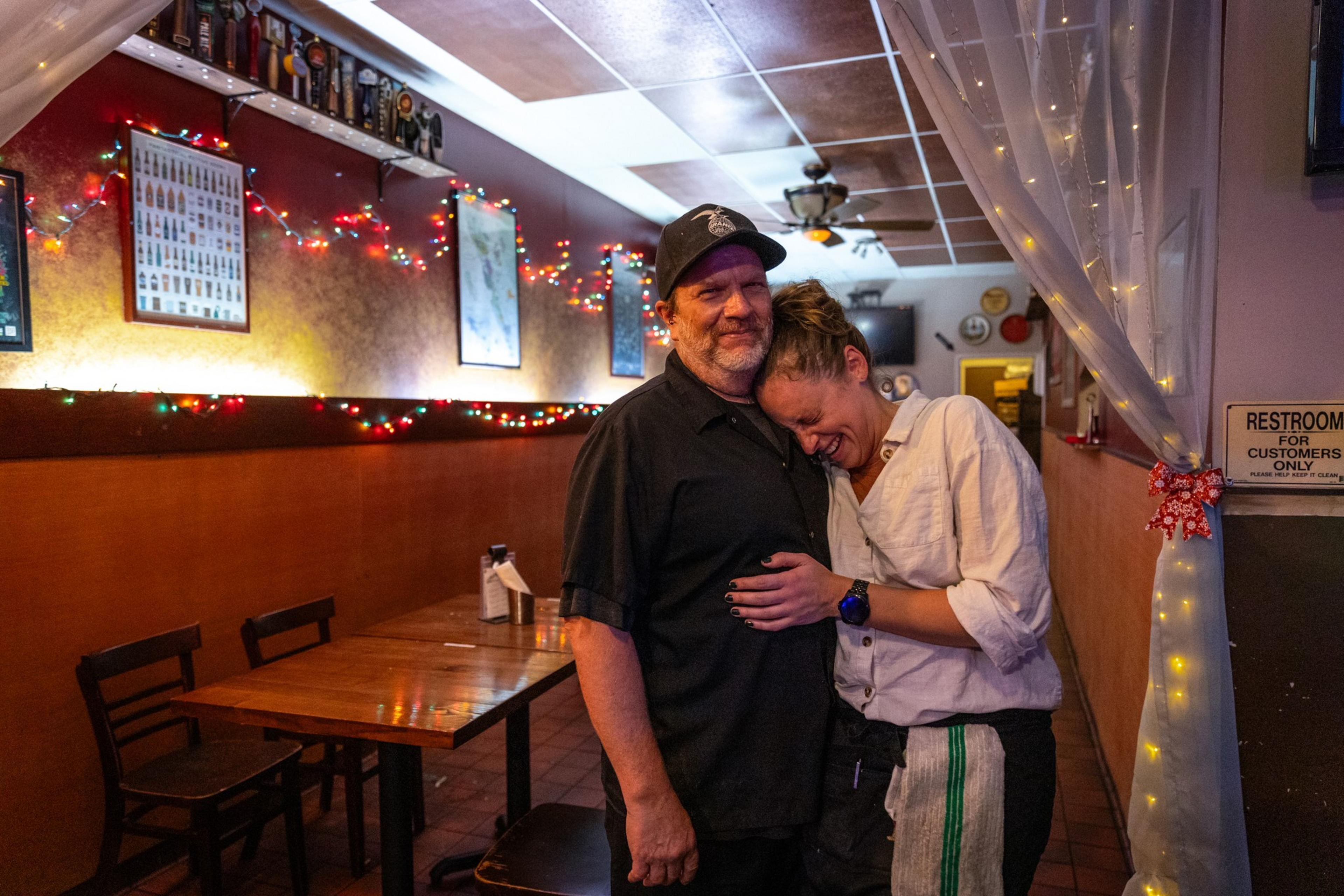 A woman hugs a man and laughs into his chest while the man looks back in to the camera smiling.