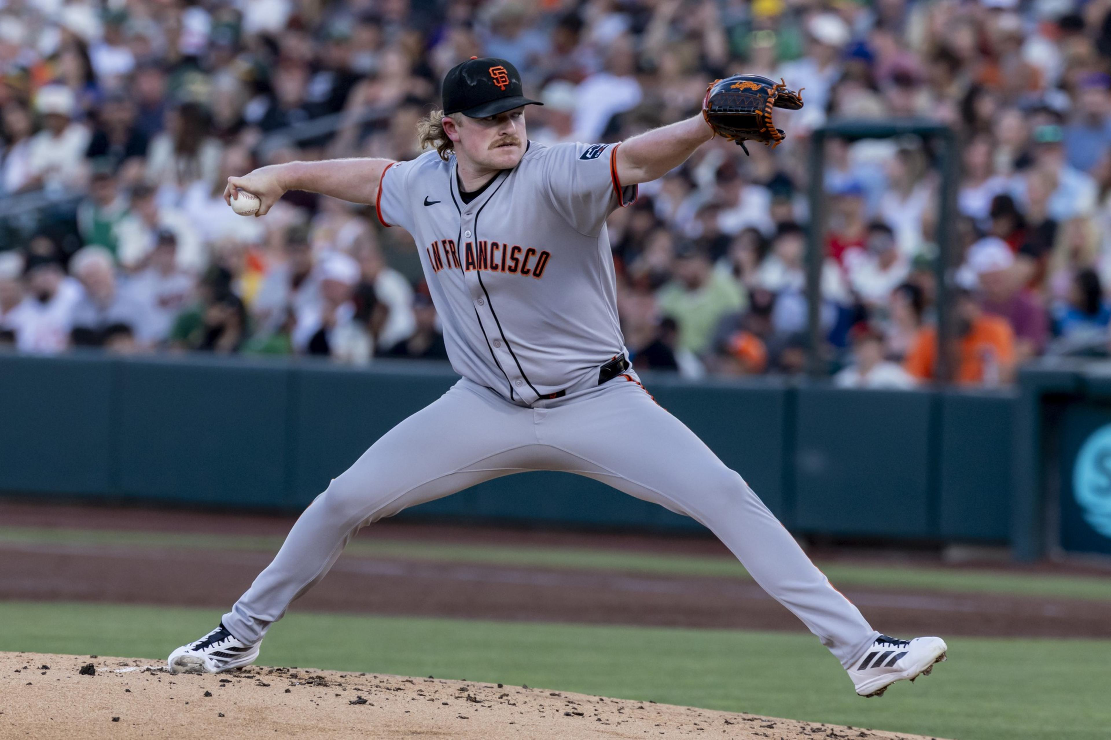 A baseball player in a grey "San Francisco" uniform pitches on a mound with a large crowd in the background. He's mid-throw, with one leg raised.