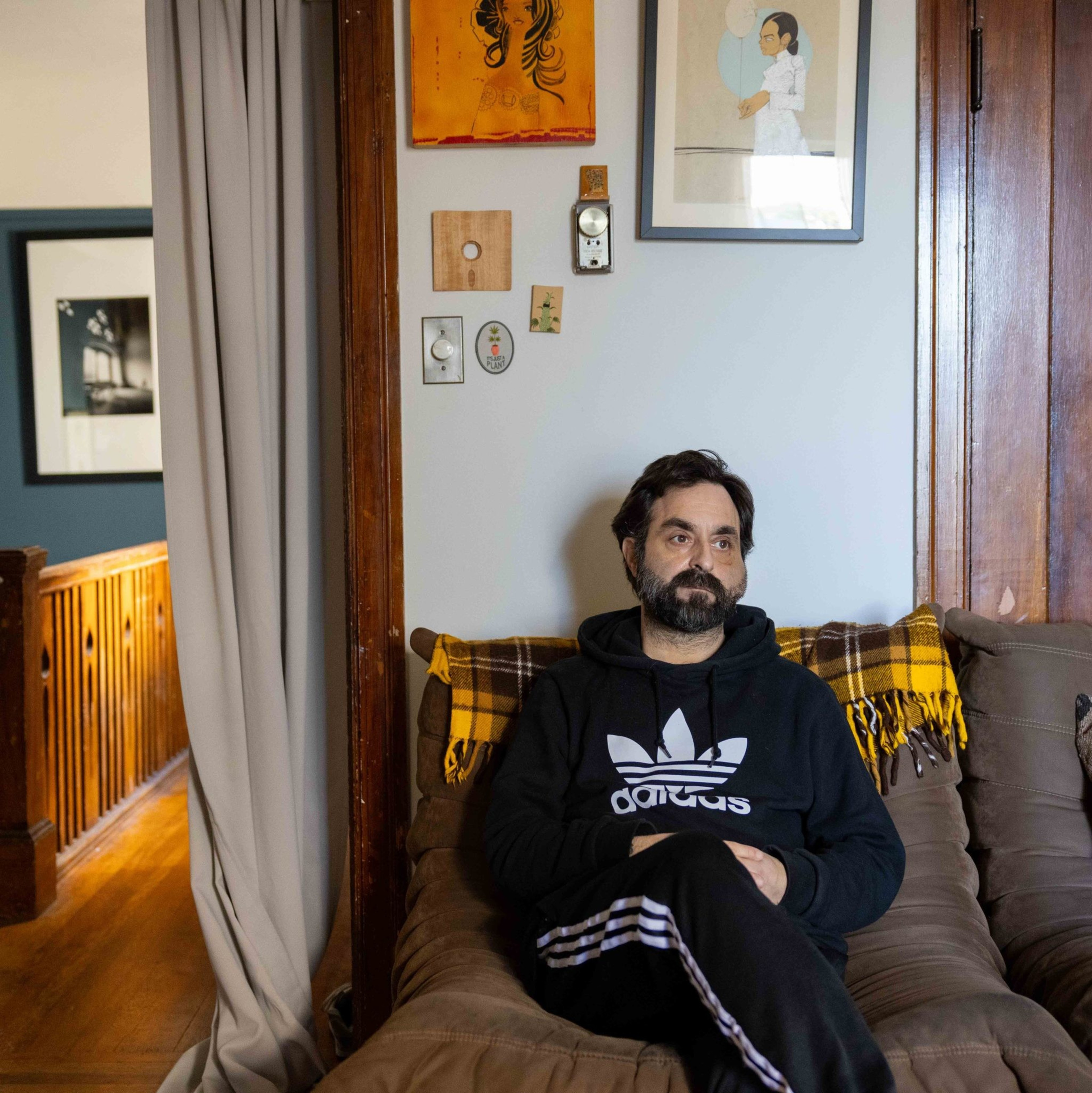 A bearded man in a black Adidas hoodie sits on a brown couch with a yellow plaid blanket. Art and photographs adorn the wall behind him.