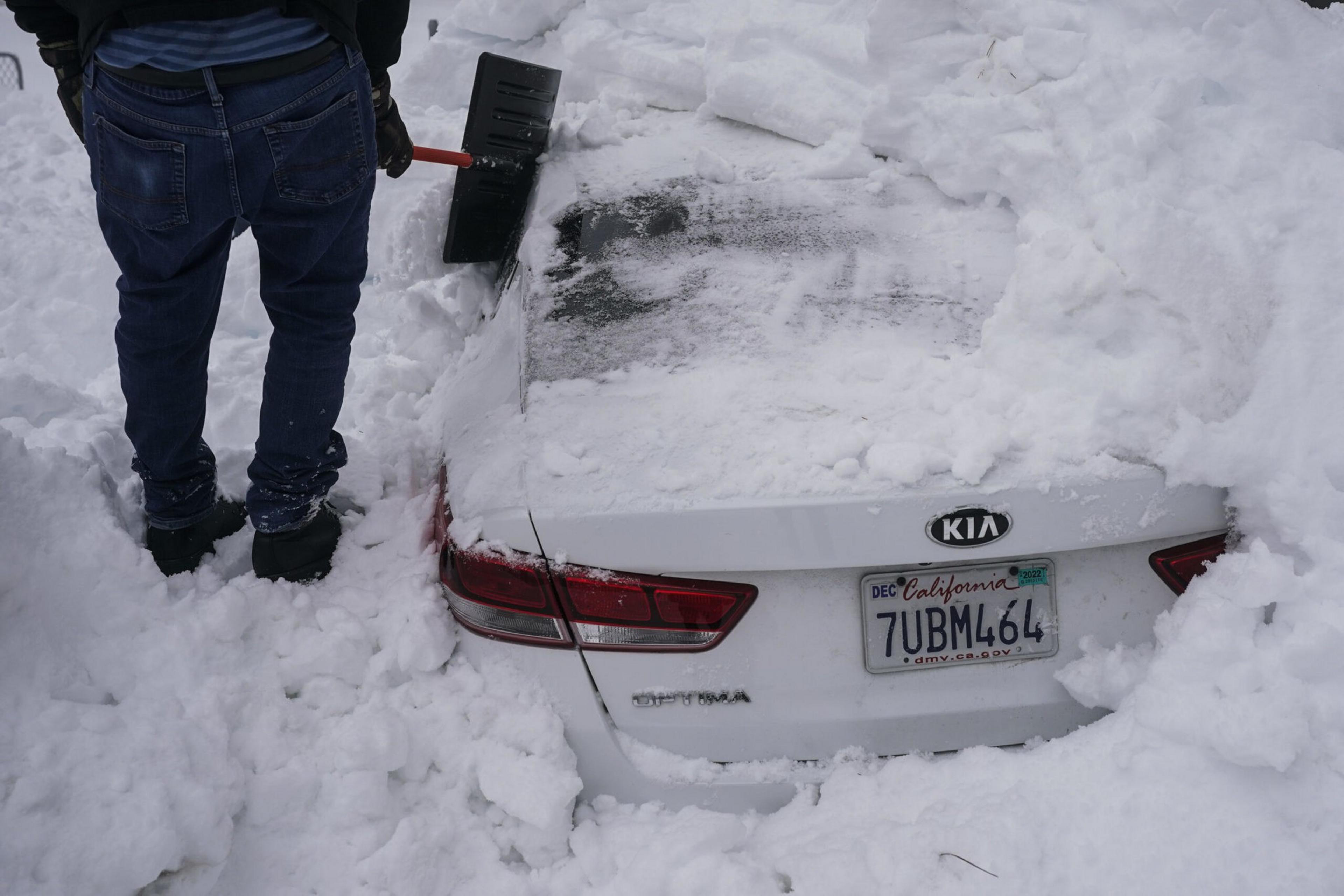 A person wearing jeans and boots is shoveling snow around a white Kia Optima car buried almost completely under thick snow.